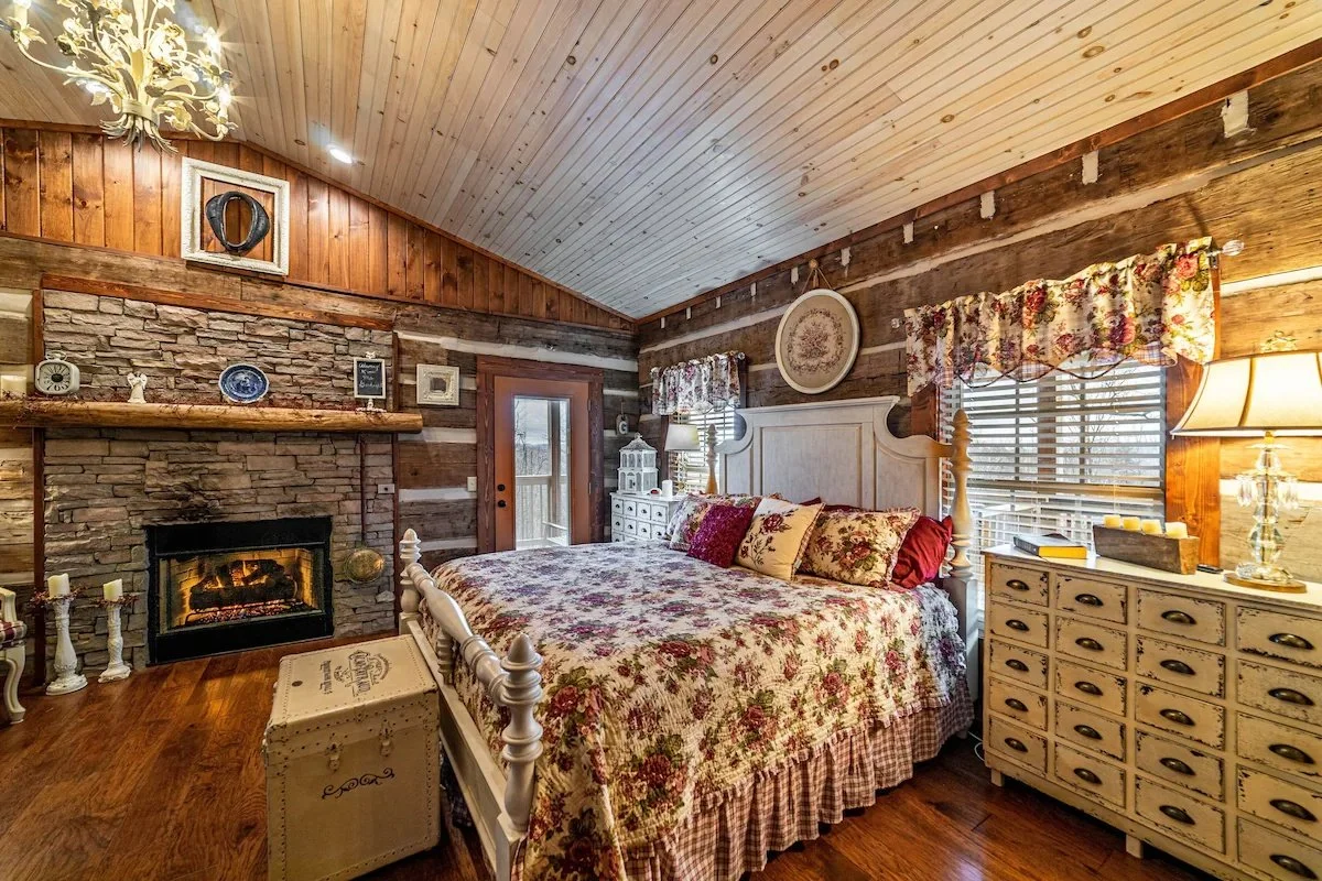 Cozy bedroom with a floral quilt, vintage dresser, and wood-paneled walls and ceiling, featuring a stone fireplace and rustic decor.