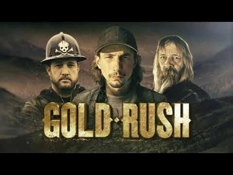 Locandina del film 'Gold Rush' con tre uomini in primo piano, uno con casco da minatore, un altro con cappello e barba, e un terzo con cappello e barba lunga, sfondo montano.