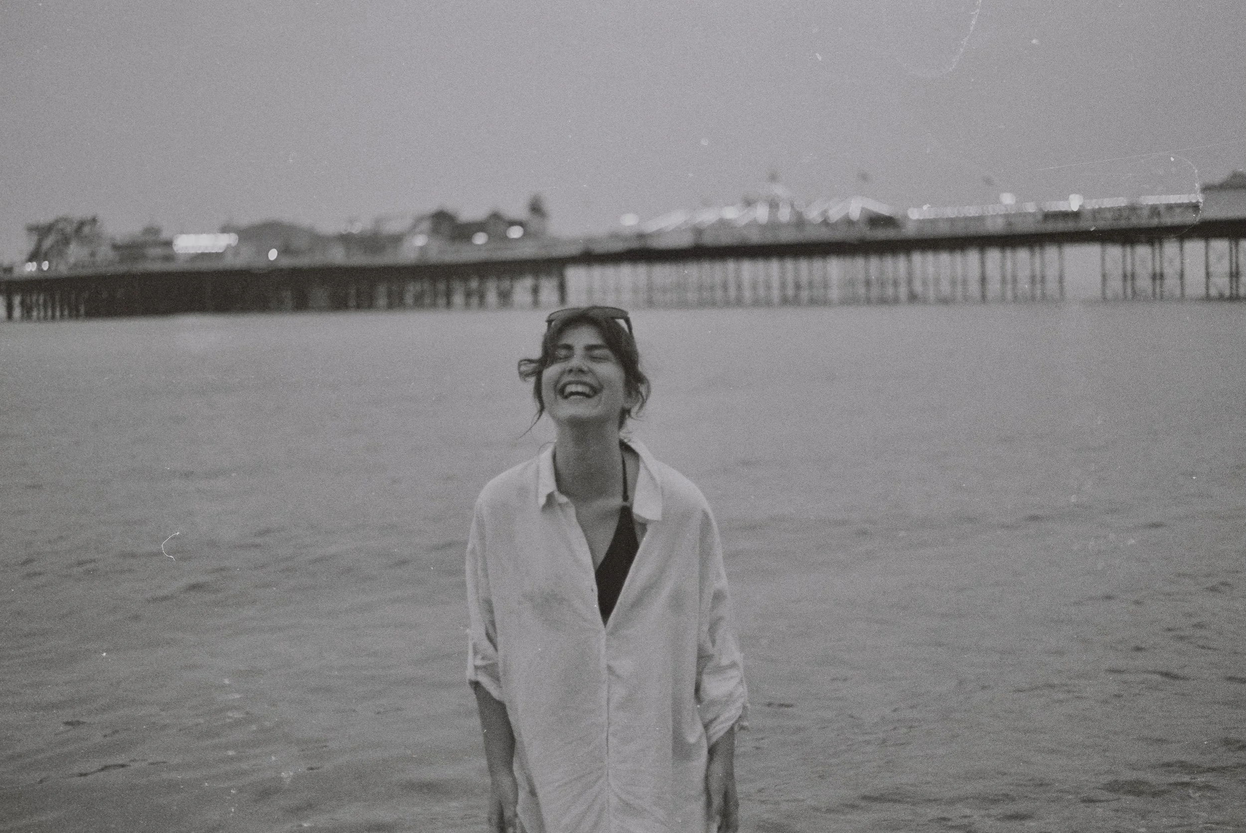 A woman smiling and walking along a beach at night, with a pier extending into the water and blurry city lights and hills in the background.