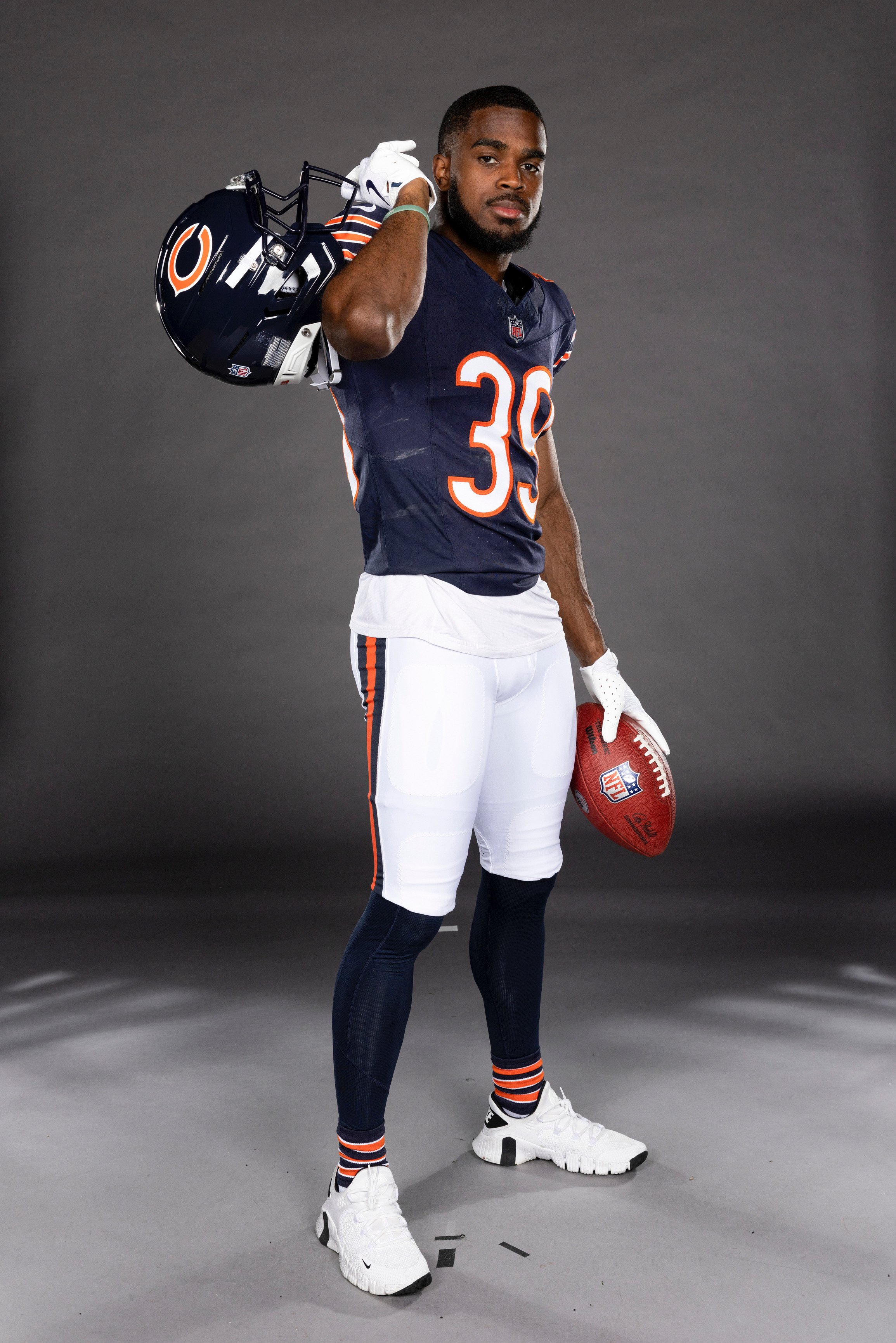 A male football player in a navy blue Chicago Bears jersey with number 39, white pants, navy and orange socks, white shoes, holding an NFL football, and carrying a helmet over his shoulder, standing against a gray background.