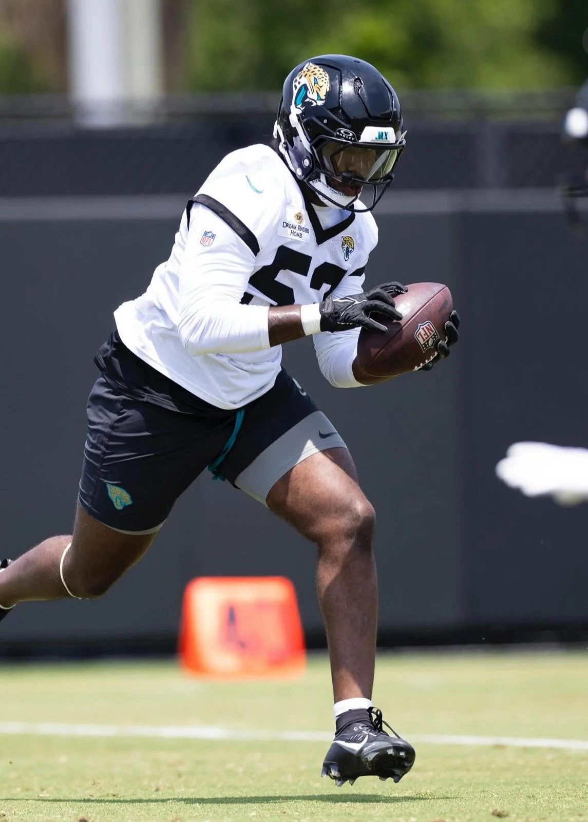 An American football player, wearing a white jersey with the number 52, black helmet with a jaguar logo, black gloves, and black shorts with a jaguar emblem, running on the field while holding a football.