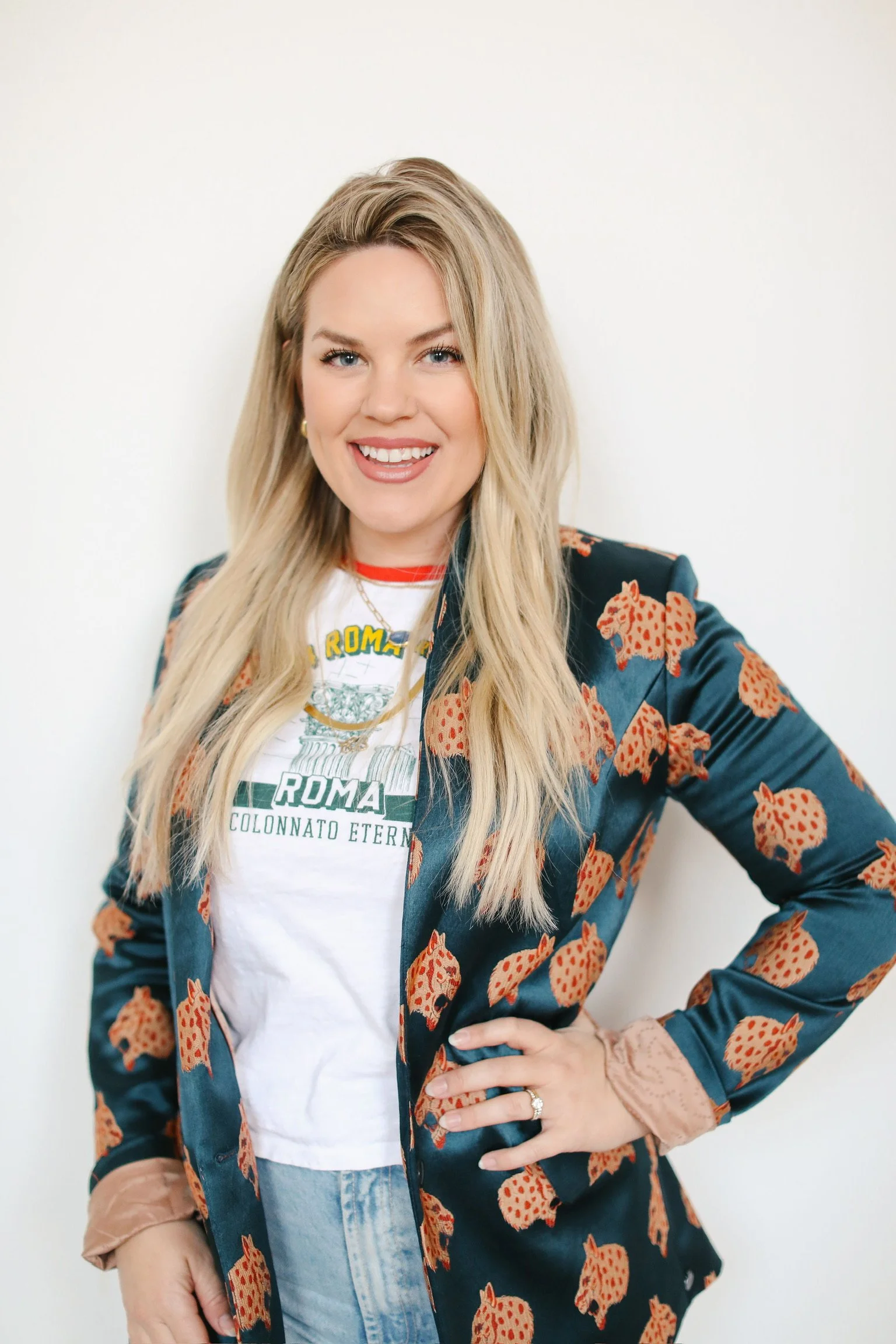 A woman with long blonde hair smiling, wearing a white graphic T-shirt, a navy blazer with orange leopard print, and light blue jeans, standing against a plain white wall.