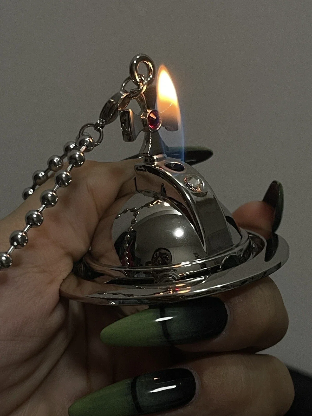 Photo of an orb lighter in silver metal activated with fire releasing. A chain is attached and out of frame. The hand holding it has wicked green finger nails.