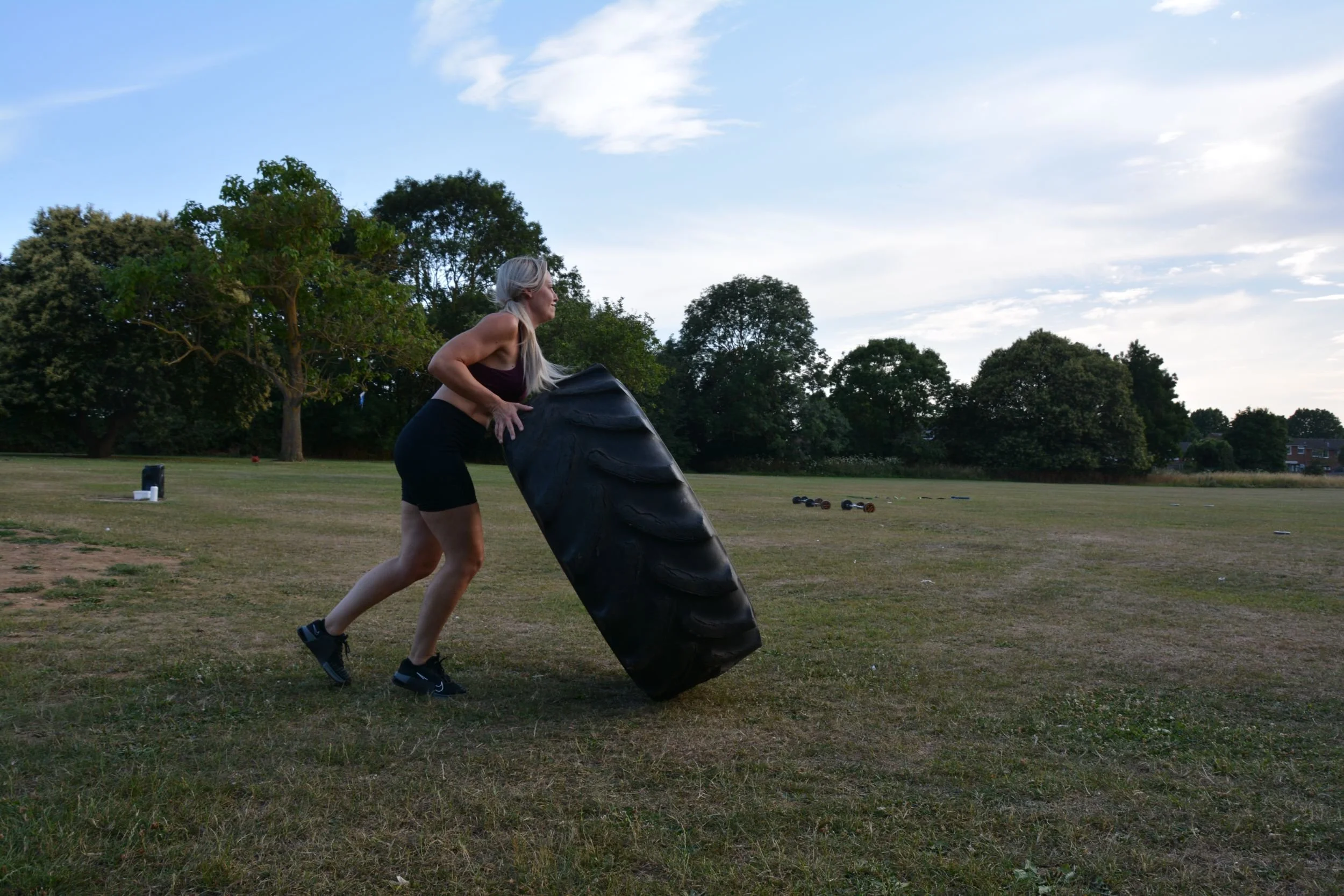 A woman is pushing a large tractor tire in an open grassy field during outdoor exercise.