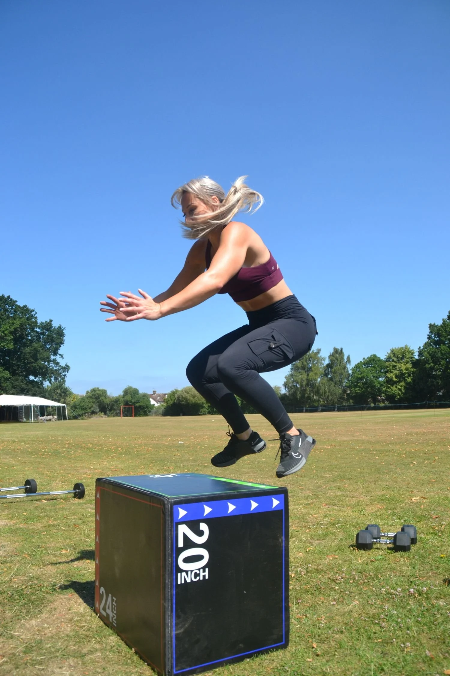 A woman jumping onto a 20-inch exercise box outdoors on a grassy field with a clear blue sky.
