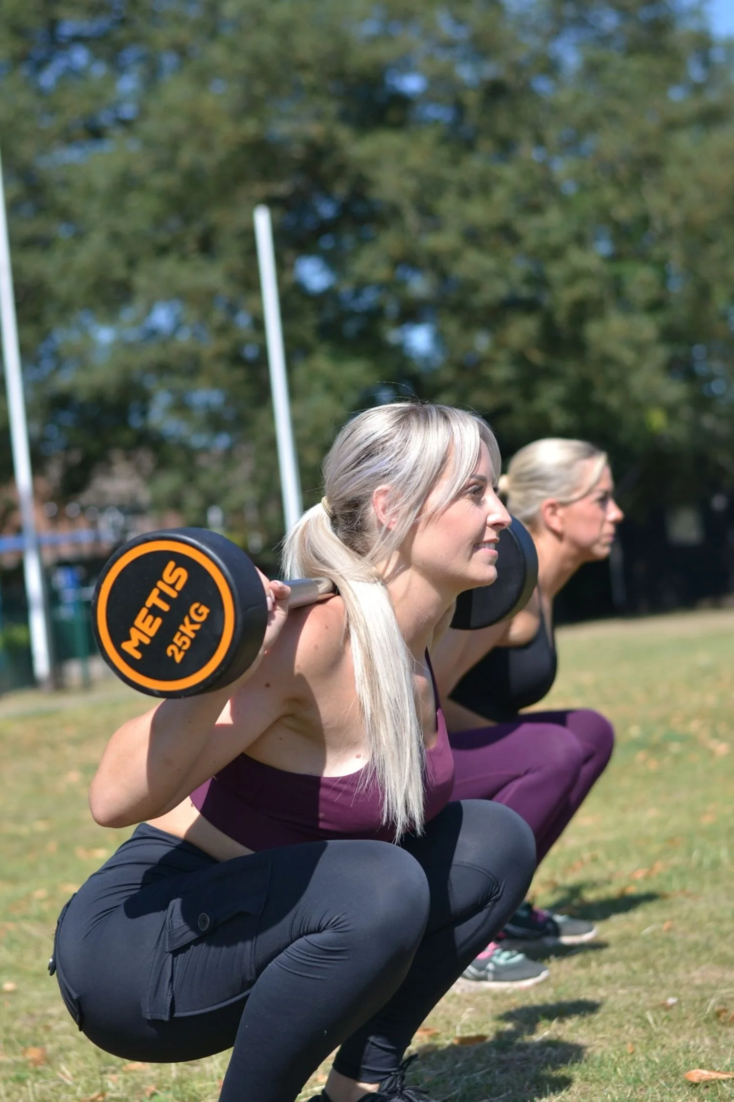 Two women with blonde hair in ponytails doing squats outdoors; the woman in front is holding a 25 kg METIS dumbbell on her shoulders.