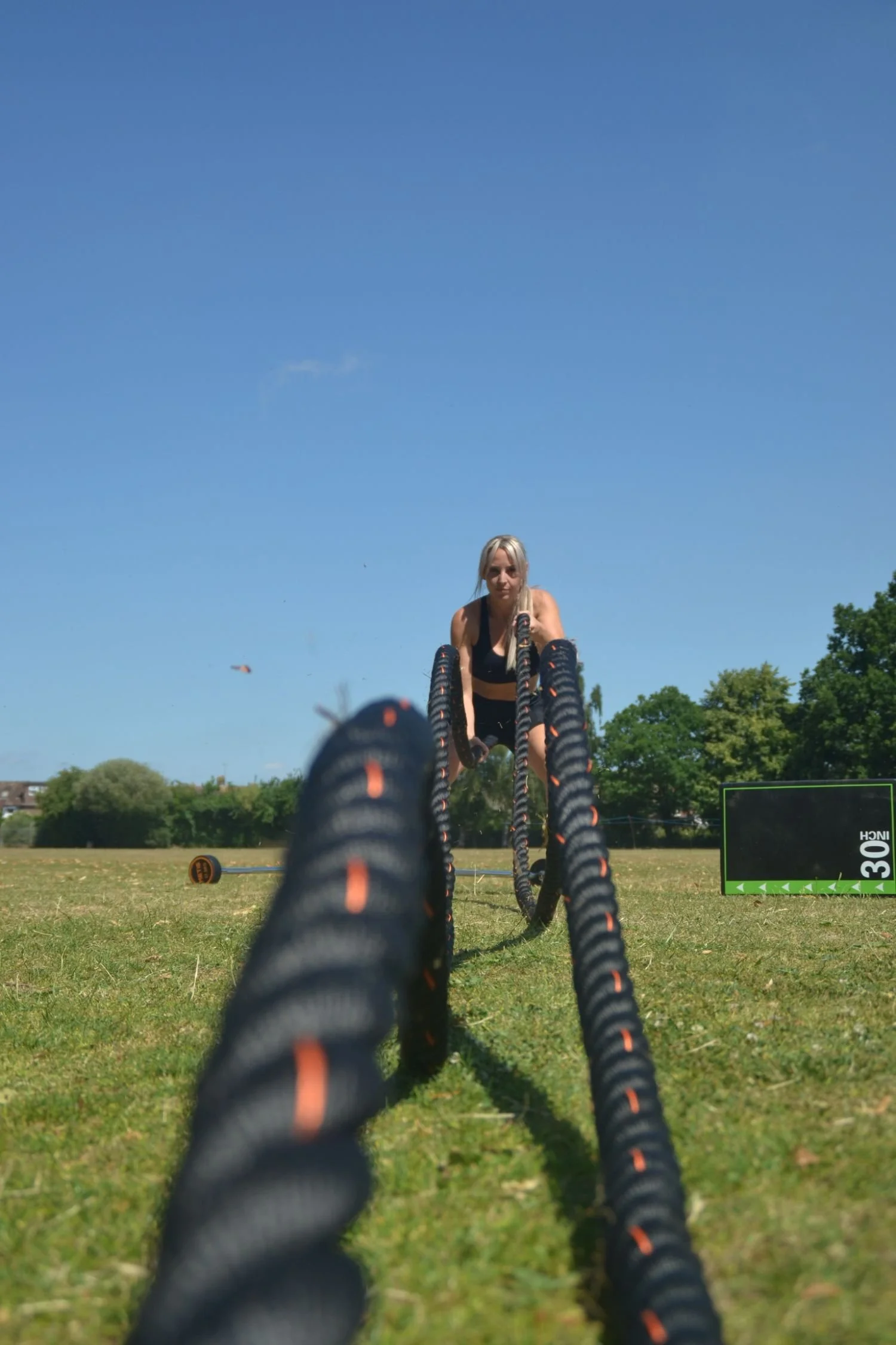 Woman using battle ropes on a grassy field during sunny weather with trees in the background.
