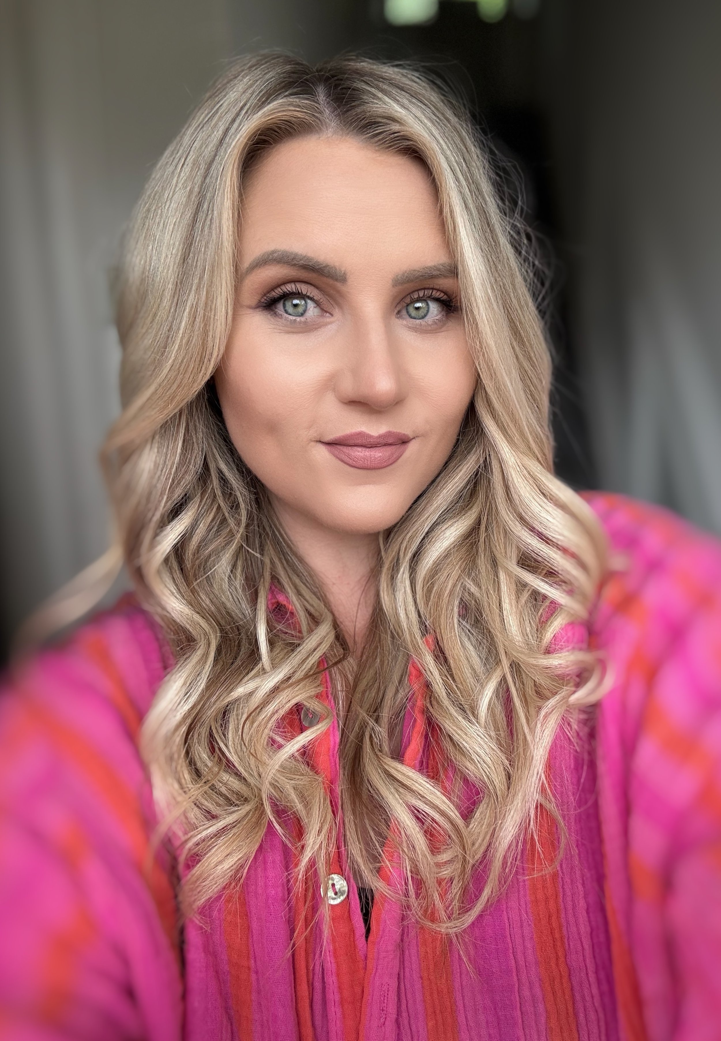 A woman with long blonde curly hair, blue eyes, and makeup, wearing a colorful pink and orange striped top, taking a selfie indoors.