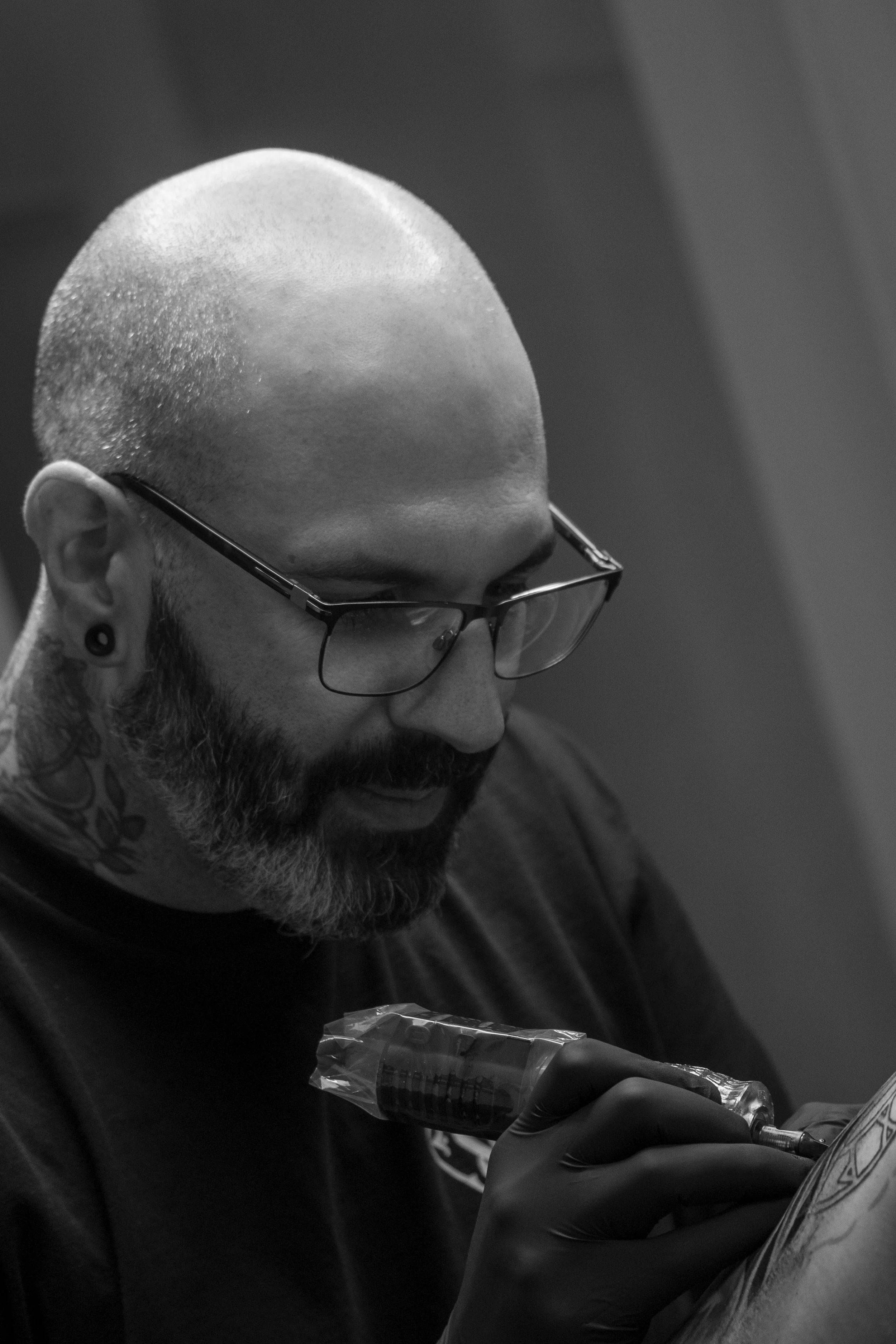 A person with glasses and tattoos is injecting a substance into their body with a syringe. They are wearing black gloves and appear to be focused on the process.