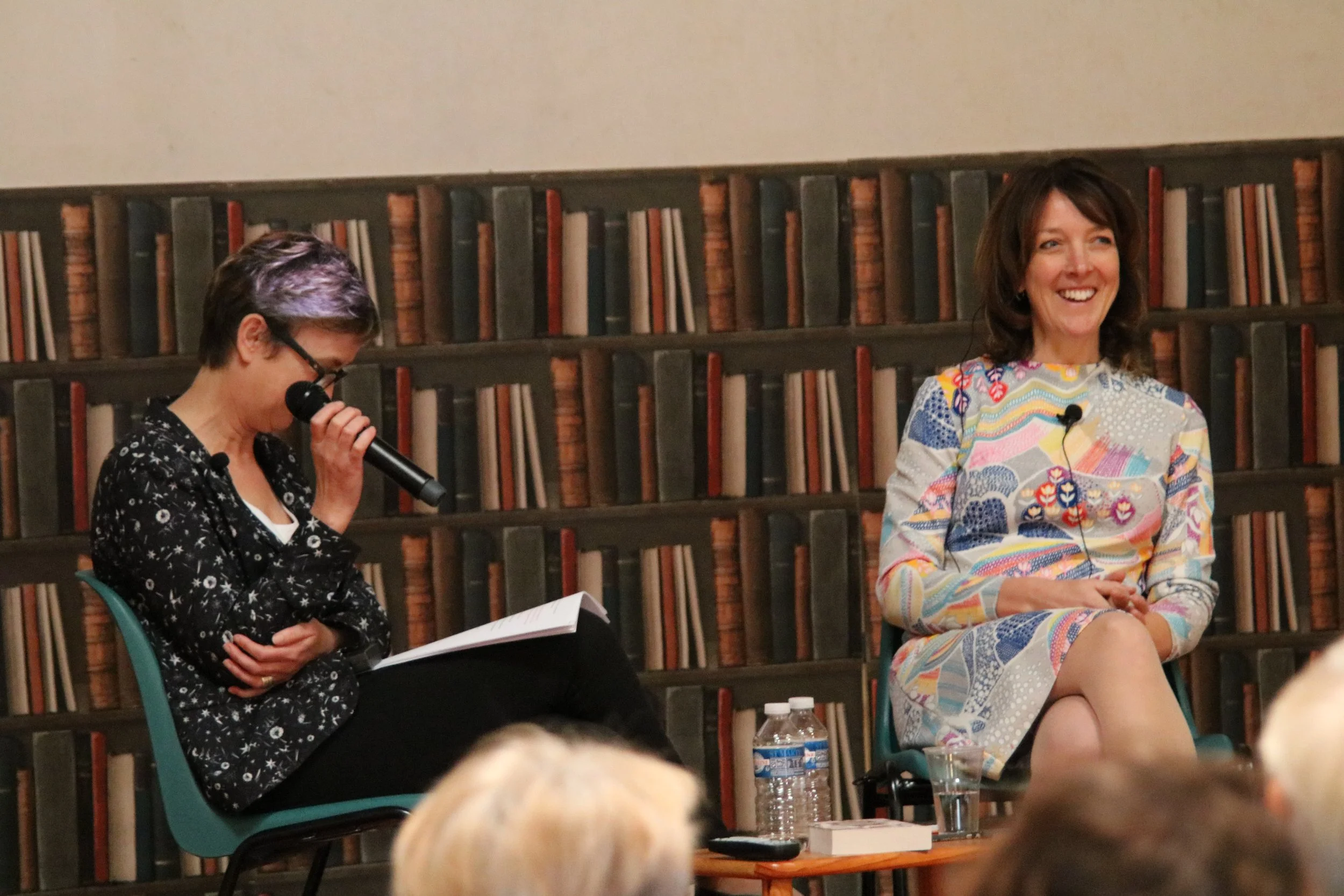 Two women seated on stage during a discussion; one woman with short dark hair and glasses, holding a microphone and a paper, the other woman with shoulder-length brown hair, smiling, wearing a colorful dress, with bookshelves in the background.