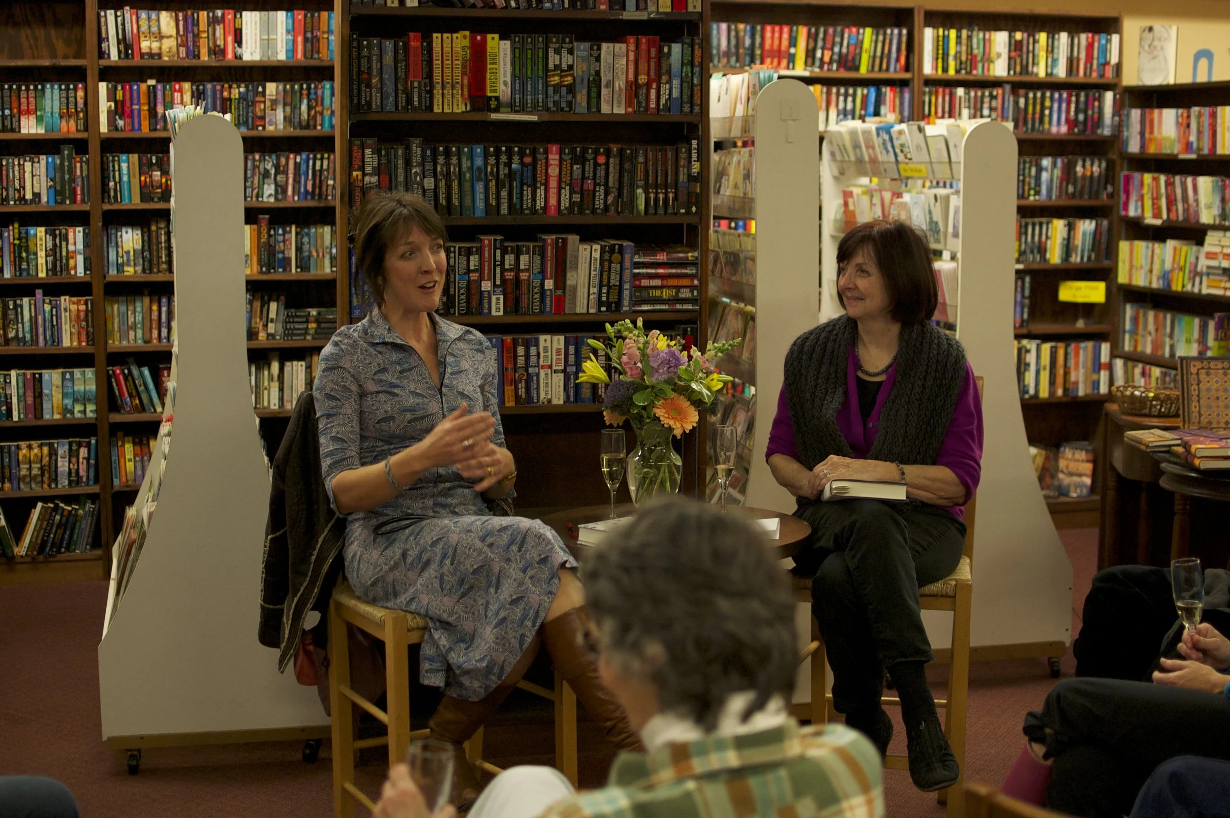 Two women are seated in a bookstore, engaged in a discussion or interview. The woman on the left is speaking animatedly, while the woman on the right listens, smiling. There is a small table with a bouquet of colorful flowers and two glasses of champ