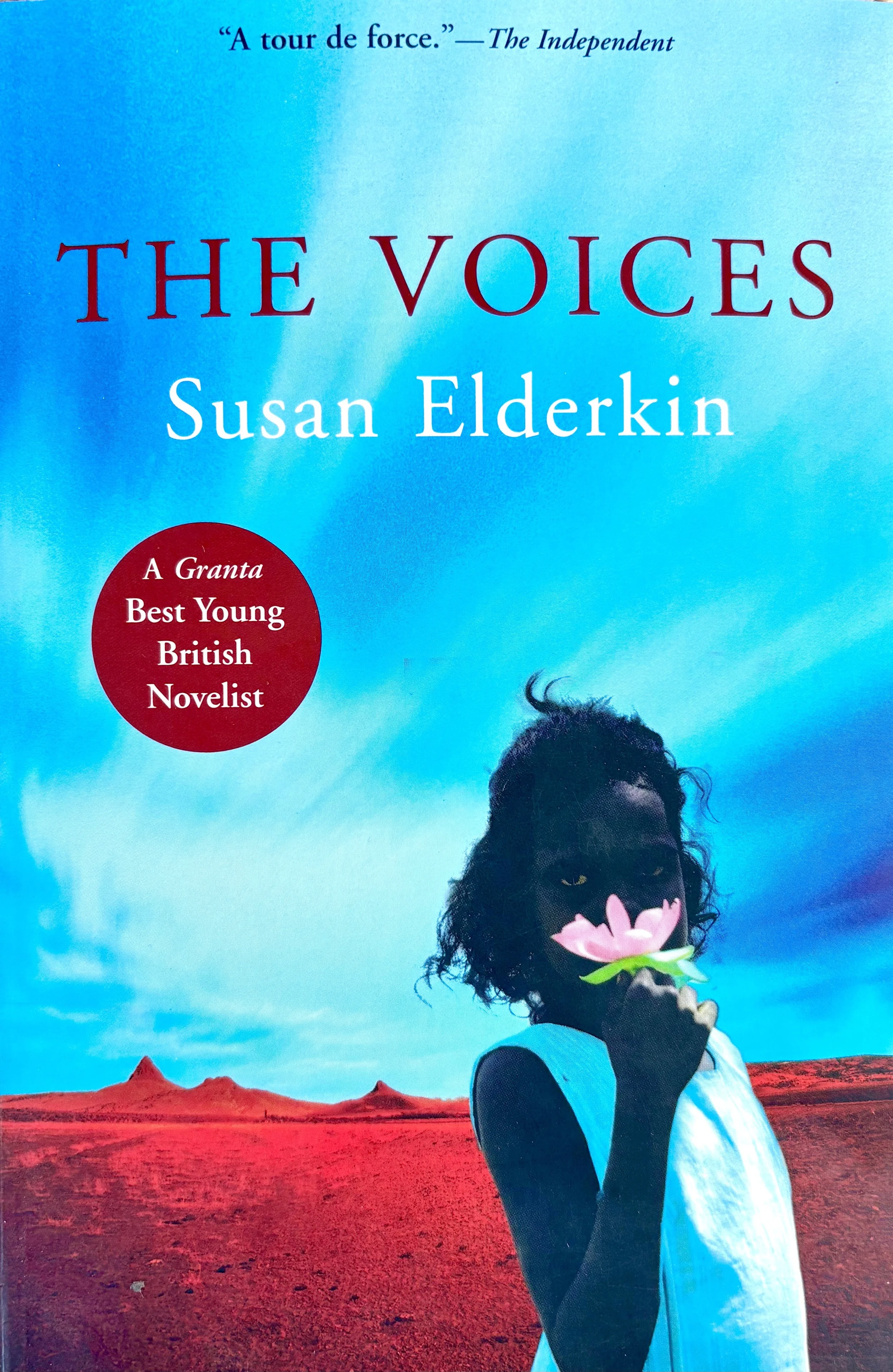 Book cover of 'The Voices' by Susan Elderkin featuring a woman holding a pink flower to her face, with a red landscape and a partly cloudy sky background.