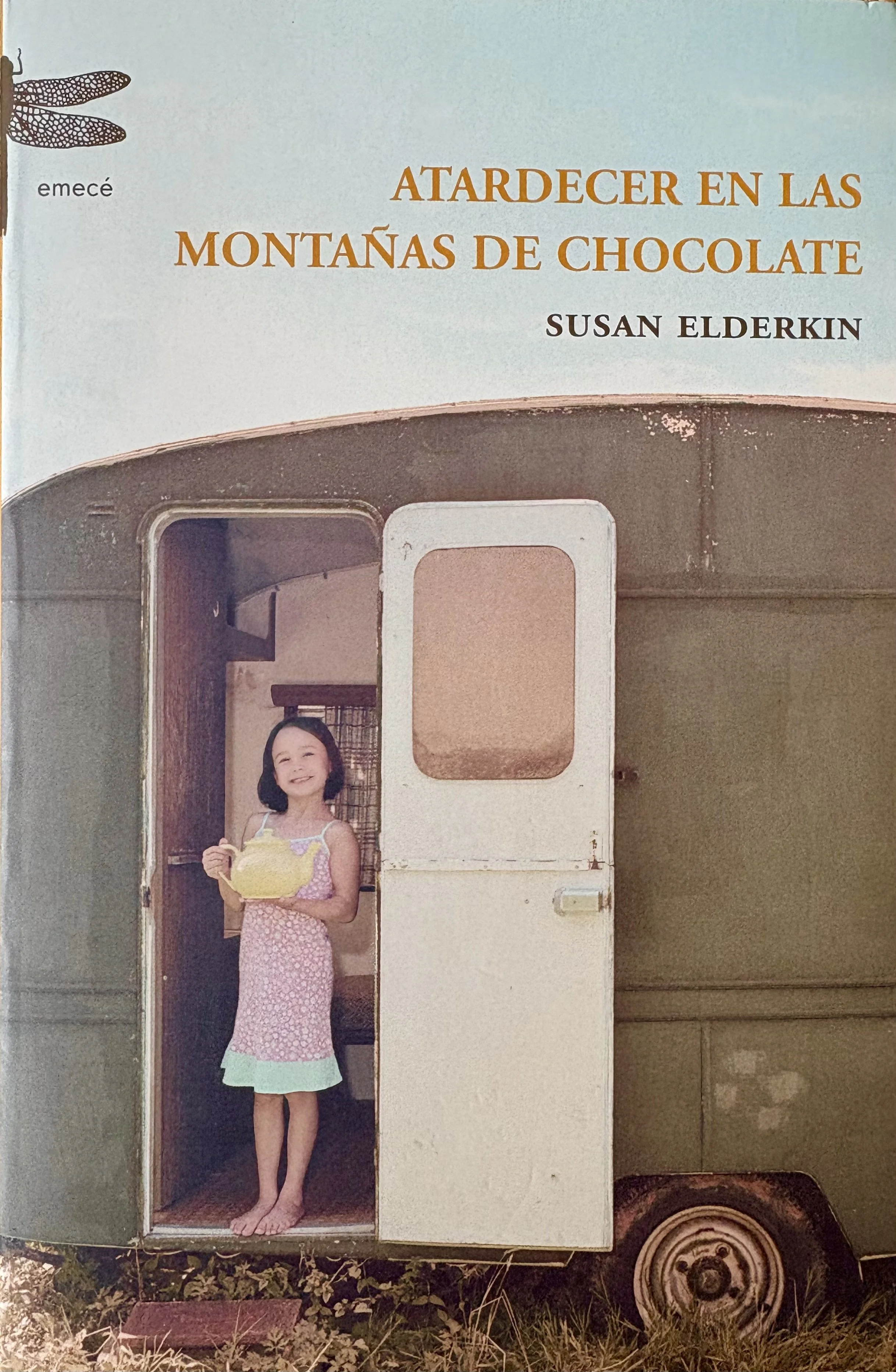 A young girl with dark hair, wearing a pink patterned dress, stands barefoot in an open door of a vintage camper trailer, holding a yellow teapot and smiling.