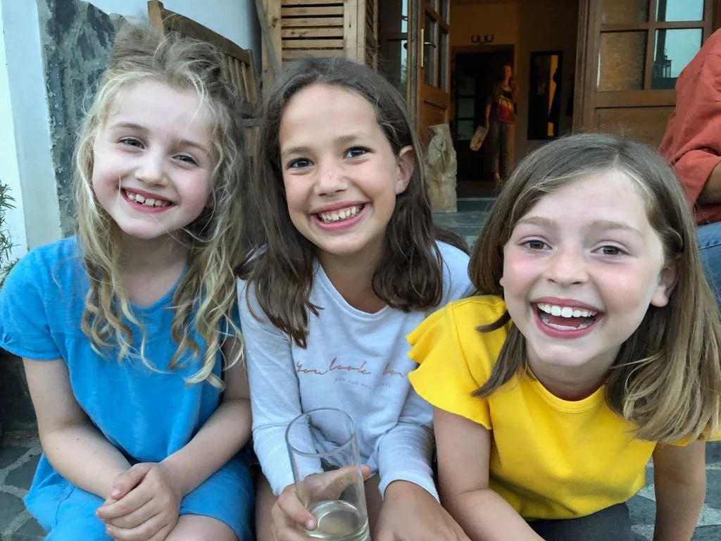 Three young girls smiling and sitting together outdoors, with two of them holding a glass of water, in front of a wooden building.