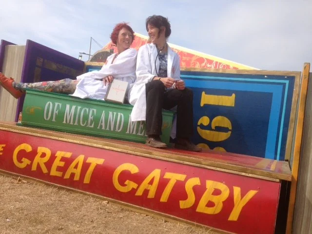 Two women sitting on a colorful float with the words 'OF MICE AND MEN' and 'THE GREAT GATSBY' displayed, at an outdoor event or parade.