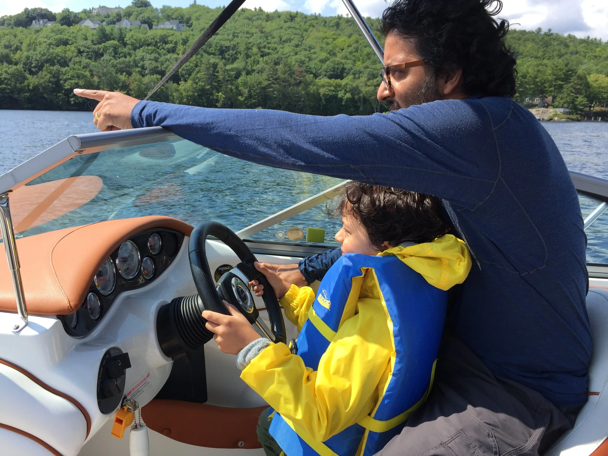 A man and a child on a boat. The man is pointing and talking, the child is holding the steering wheel. The boat is on a lake with green trees and hills in the background.