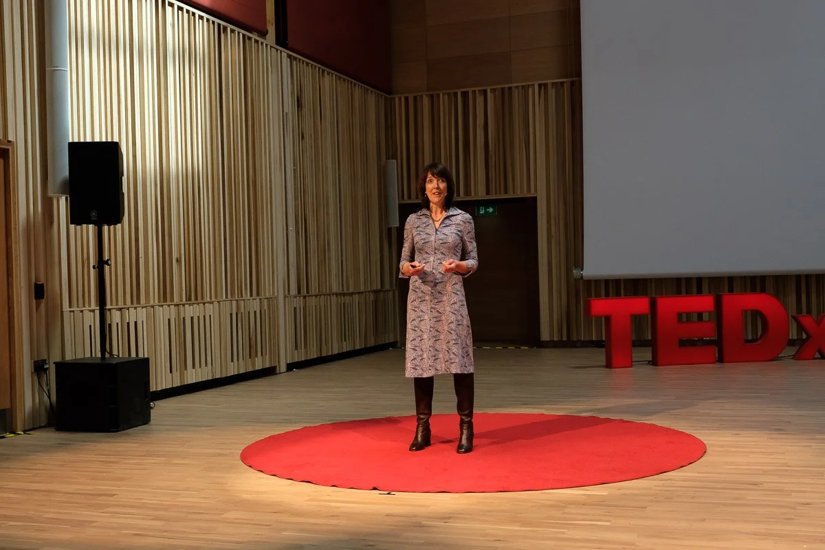 A woman giving a TED talk on a stage with a red circular carpet, large TEDx letters, and a projection screen behind her in a modern auditorium.
