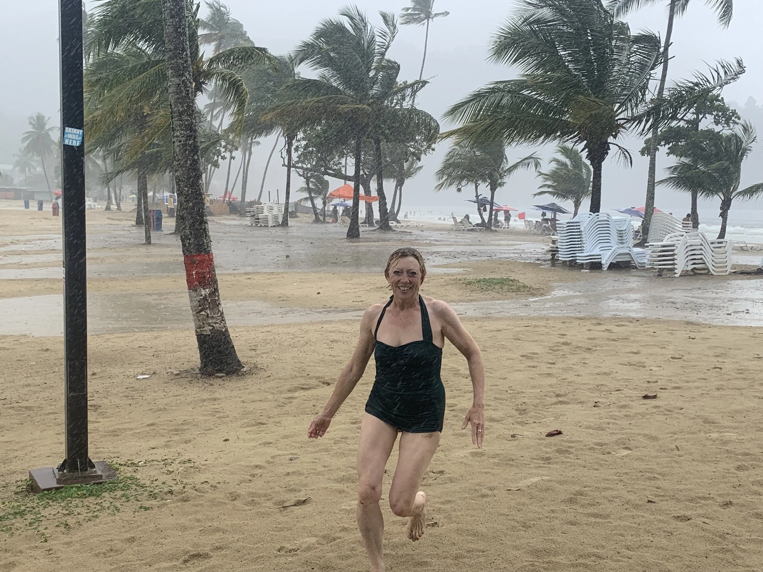 A woman in a black swimsuit smiling and playing in the rain on a sandy beach, with palm trees, umbrellas, and stacked chairs in the background.