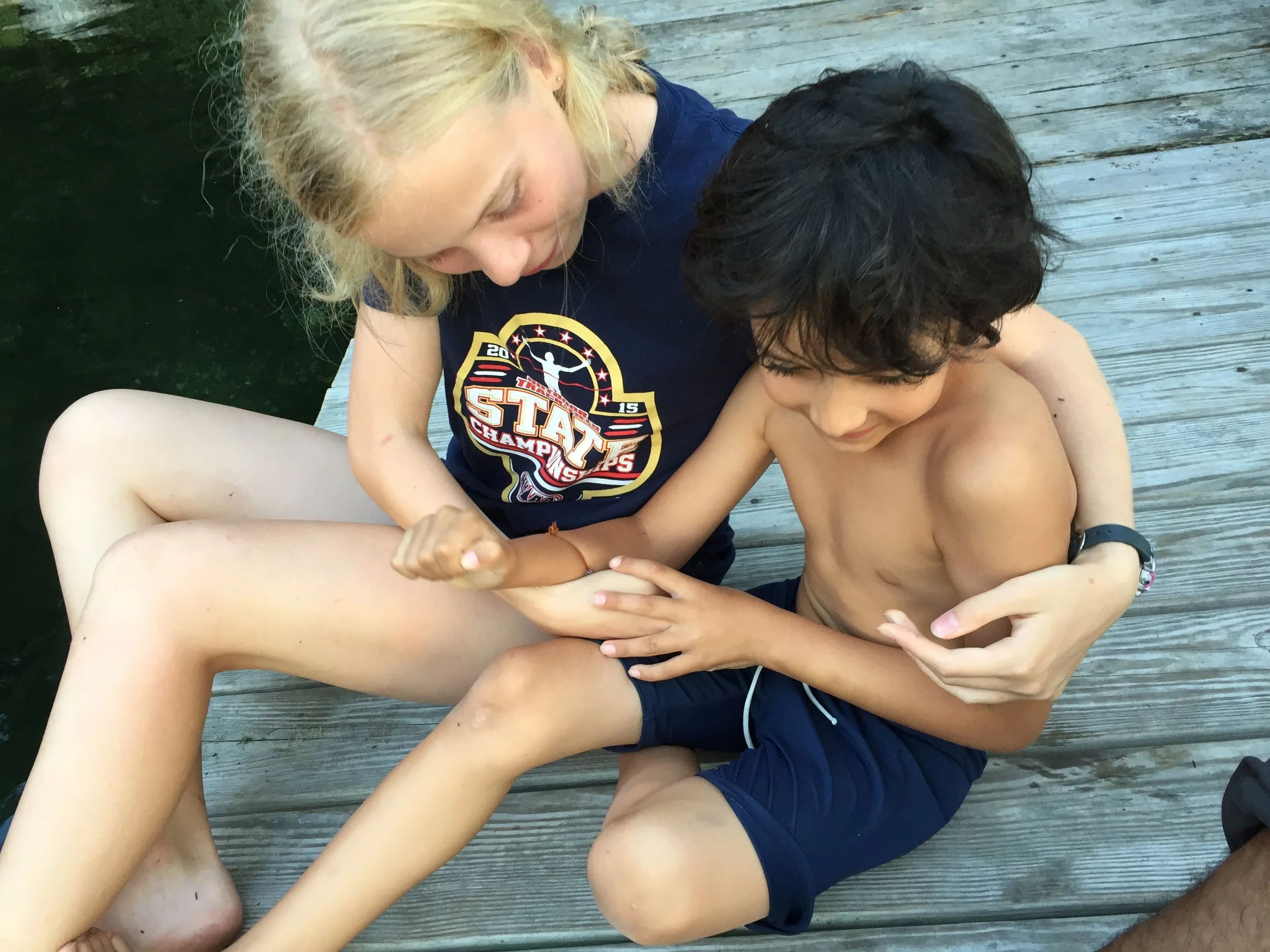 Two children, a girl and a boy, sitting on a wooden dock near water, engaging in a playful arm wrestle.