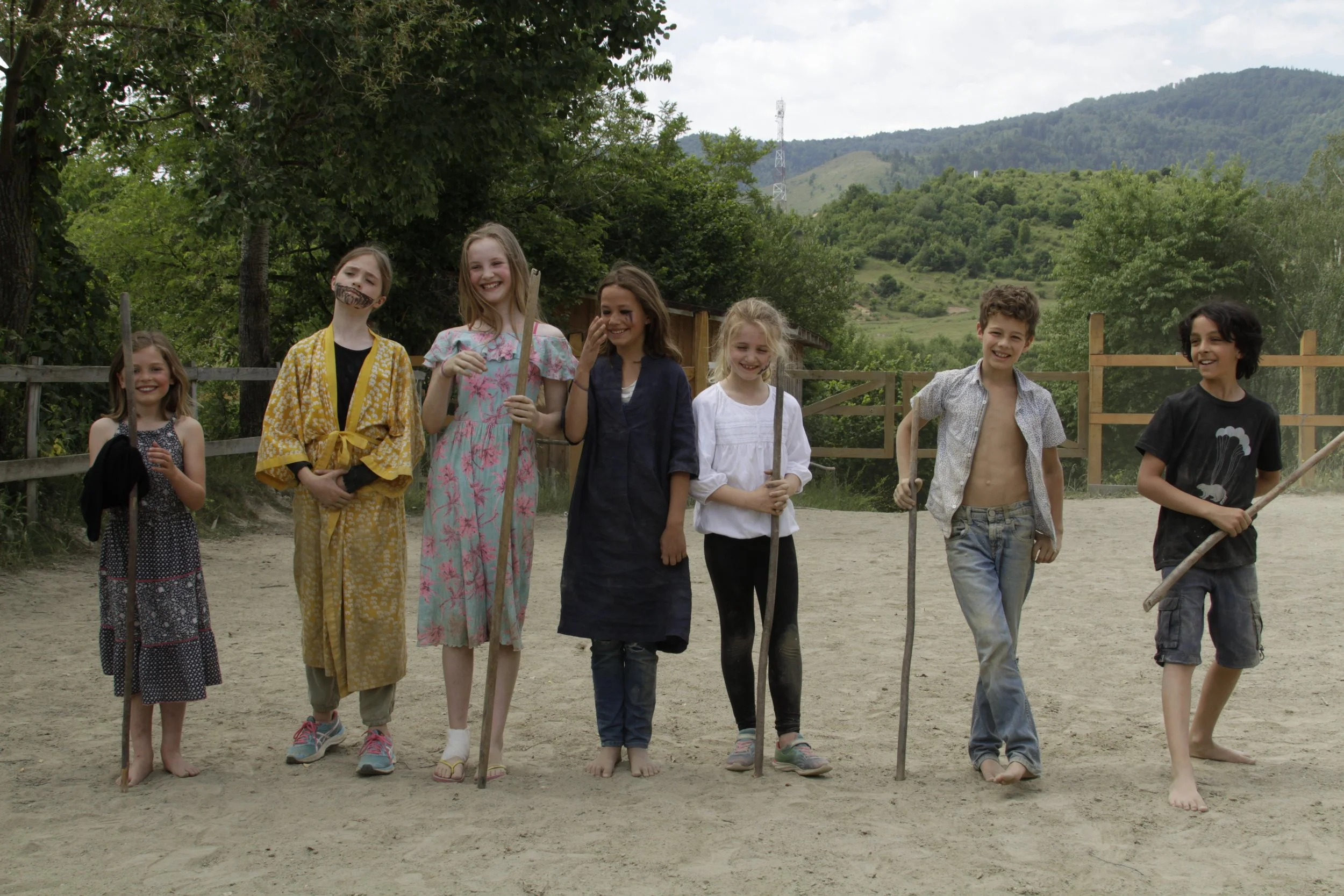 A group of eight children standing outdoors on a dirt ground, smiling and holding sticks, with trees and hills in the background.