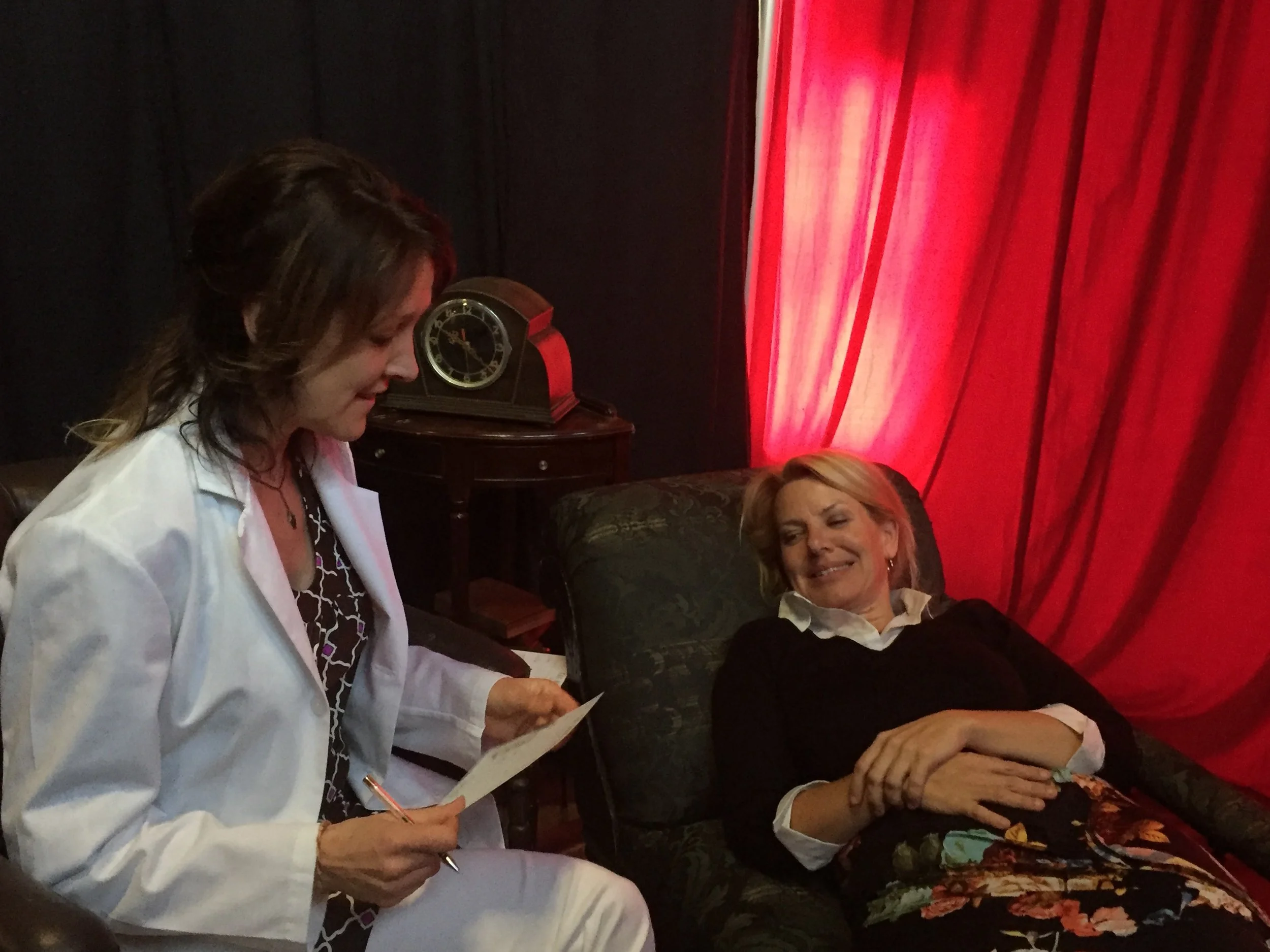 A woman lies on a sofa, smiling at another woman in a white coat who is sitting beside her and reading from a piece of paper. The room has dark walls, red curtains, and a vintage clock on a side table.