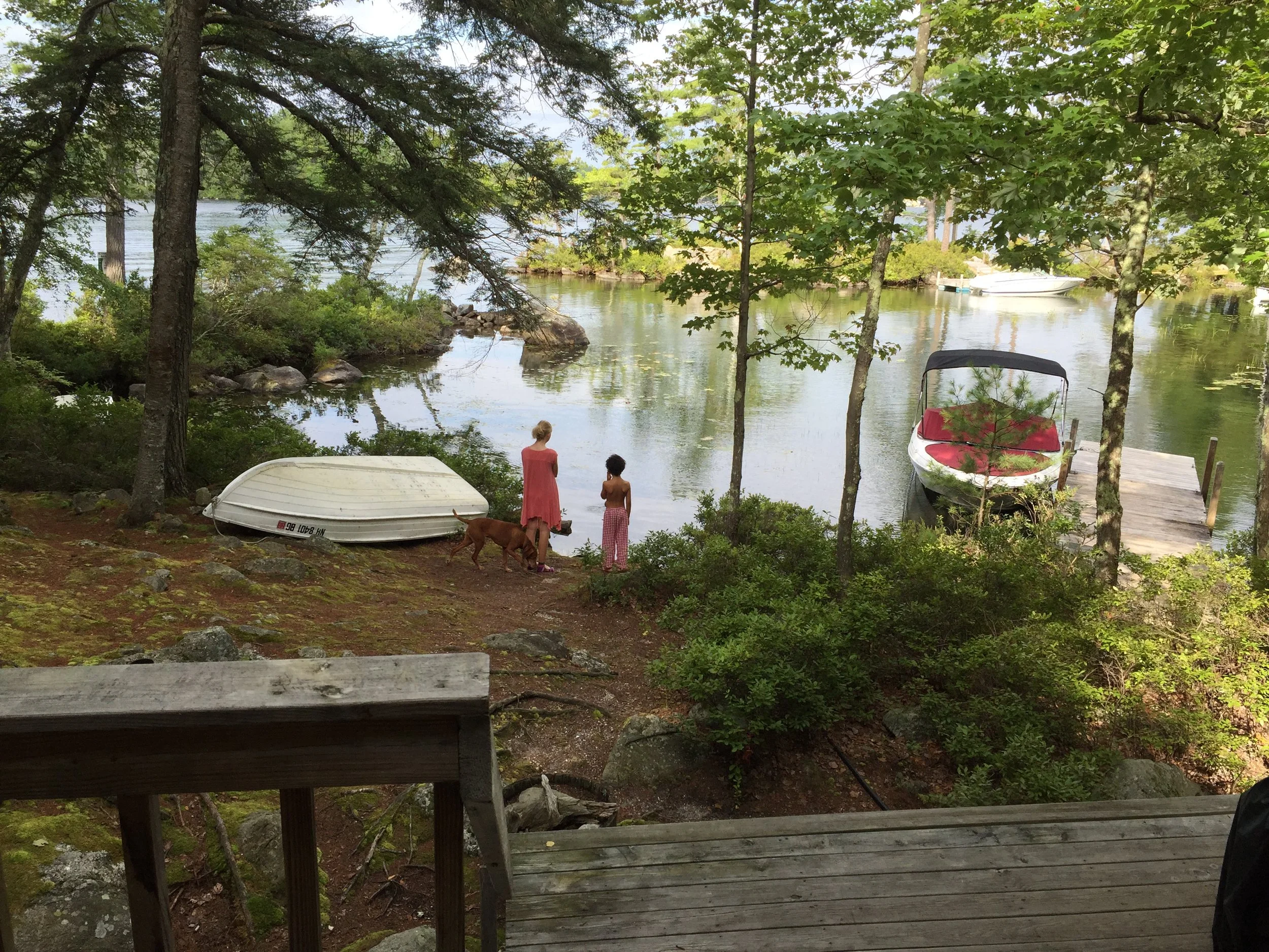 Two women and a dog by a river, with boats on the water and trees surrounding the area.