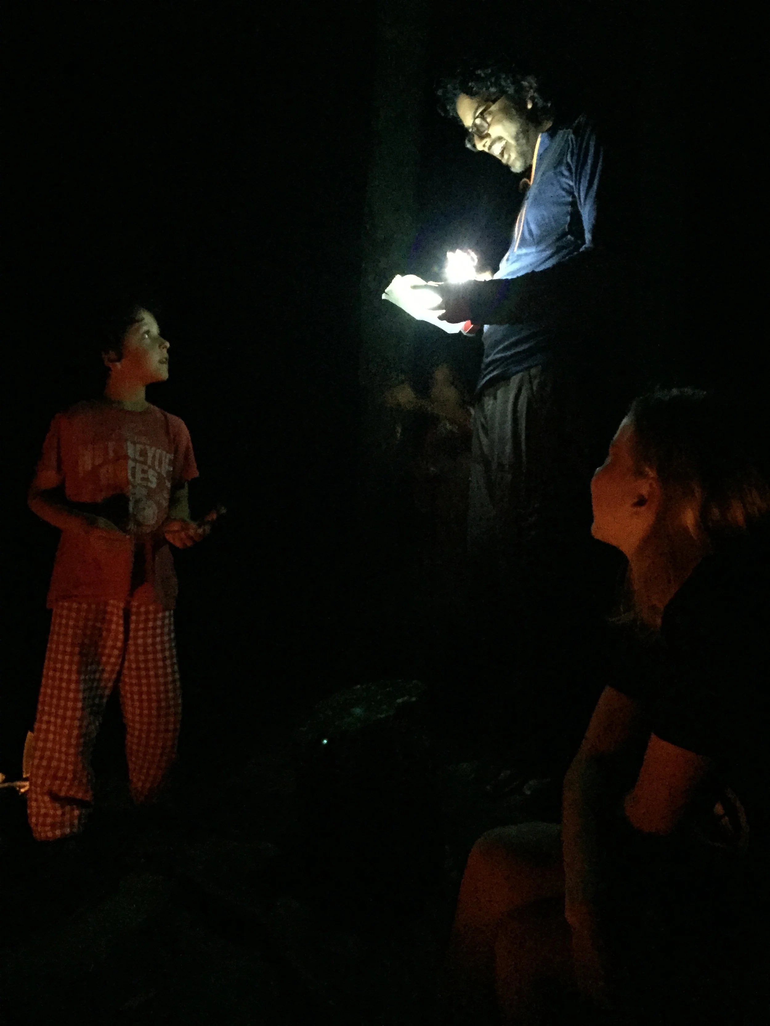 A person with glasses and curly hair, wearing a dark jacket, is holding a glowing object and smiling. Two children are looking at them; one child stands while the other is sitting, and both are in a dark environment.