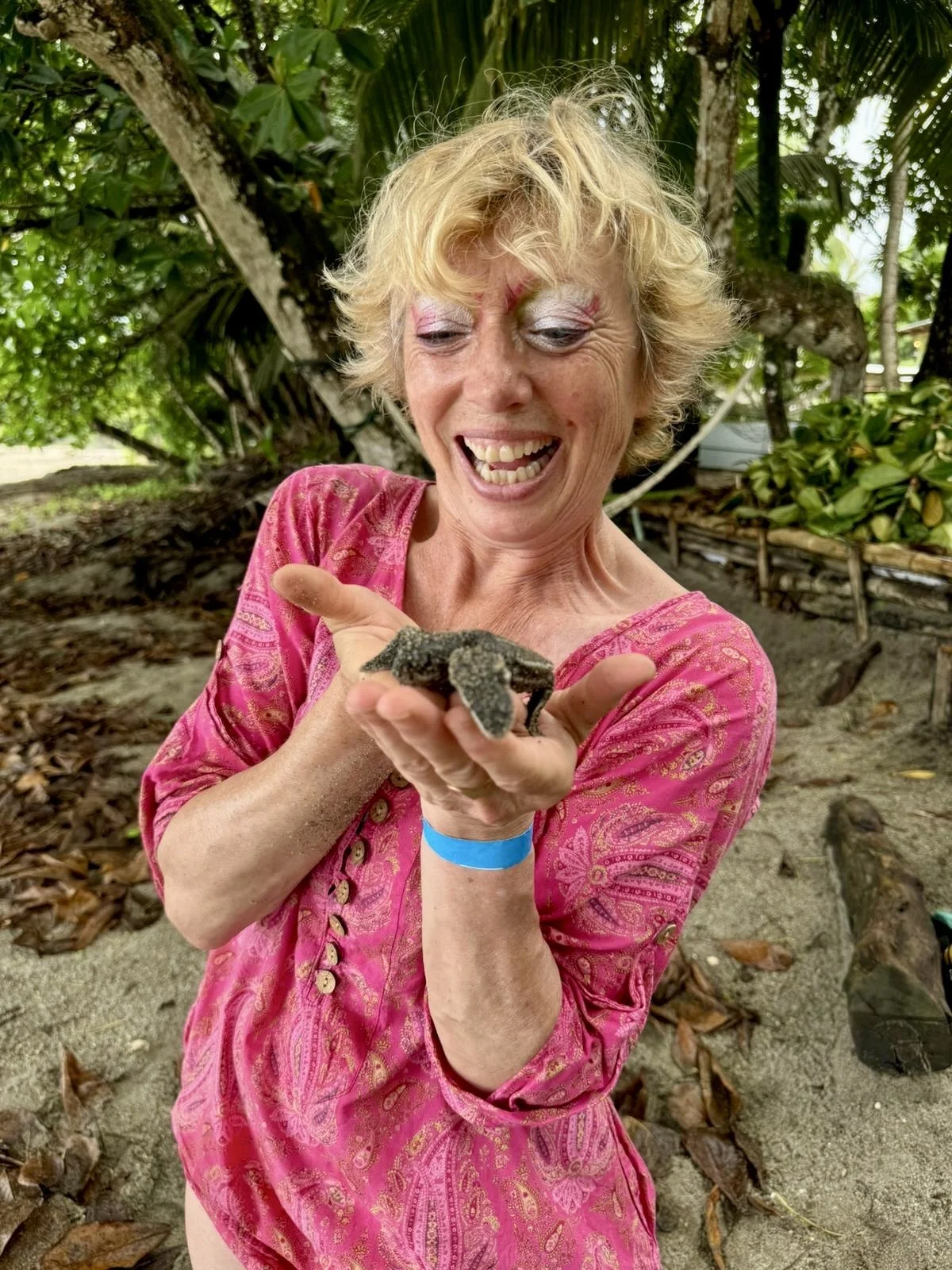 A woman with blonde curly hair in a pink dress holding a small black iguana and smiling outdoors on a sandy area with trees and foliage in the background.