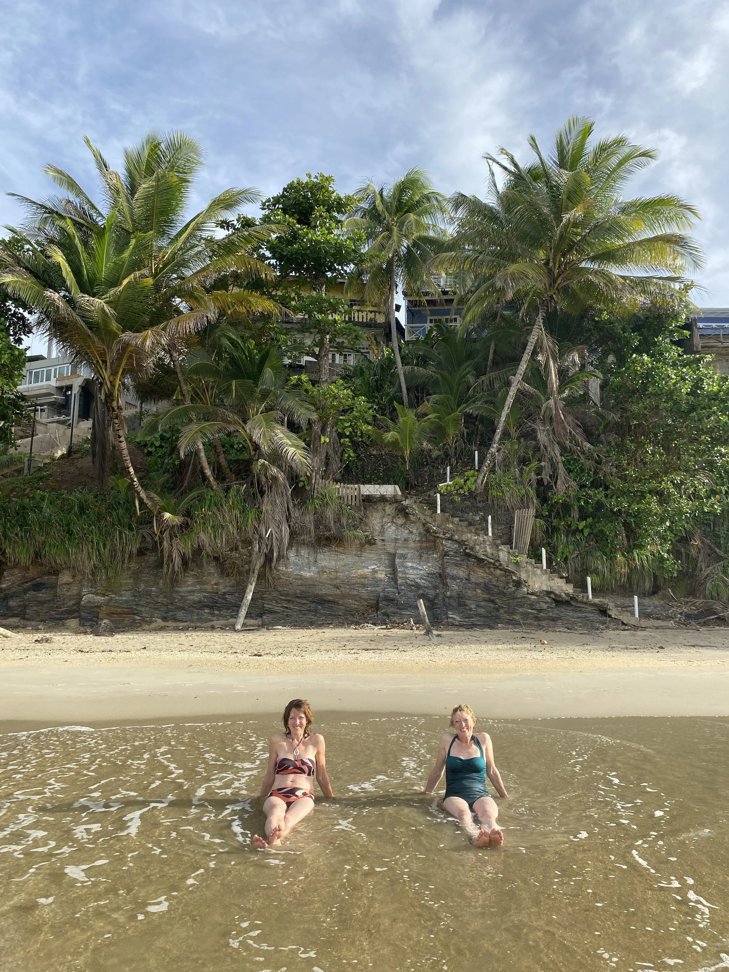 Two women sitting in shallow ocean water near the beach with palm trees and houses on a hill in the background.