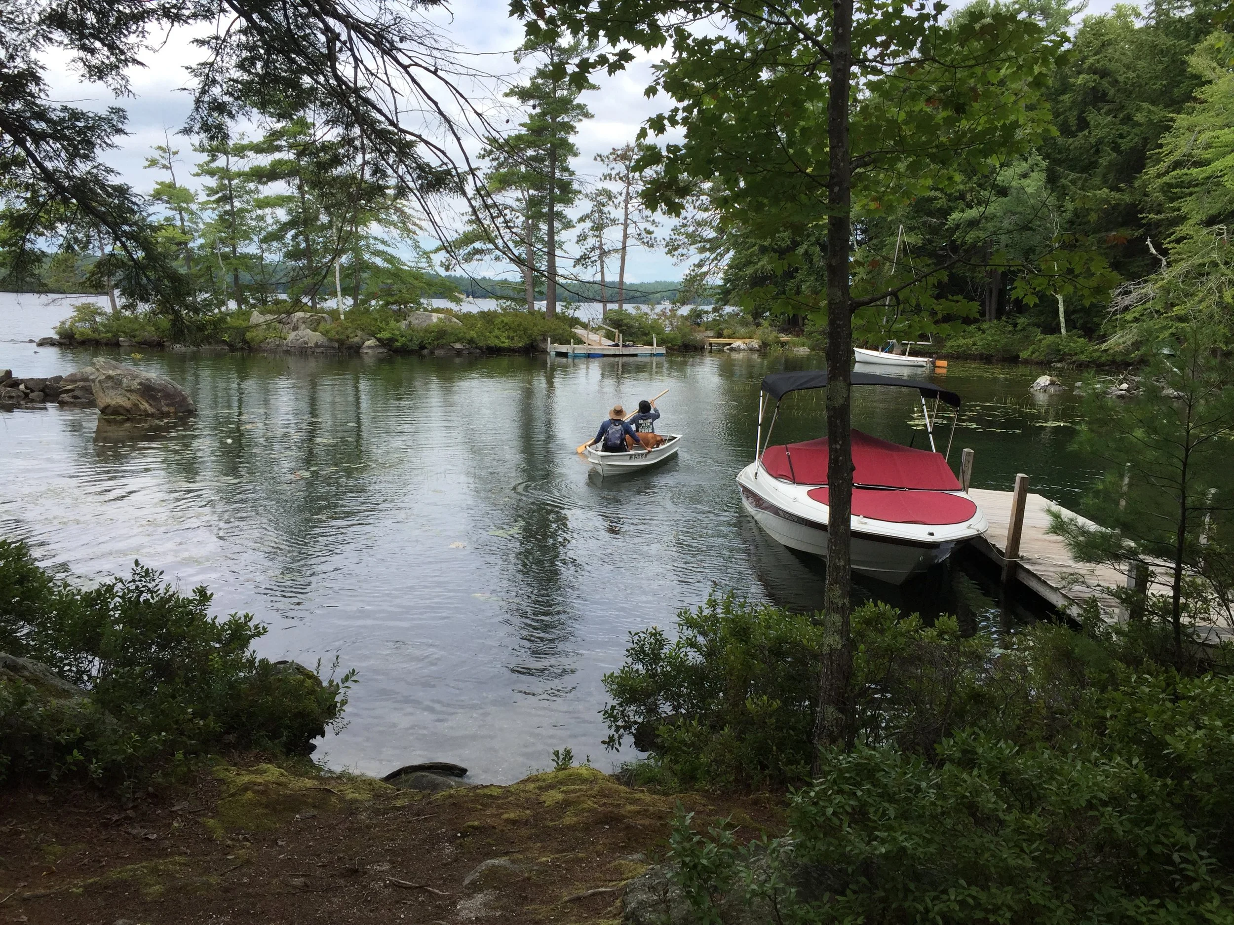 A lakeside scene with a small dock, a boat with a red cover, and two people paddling in a canoe surrounded by trees and rocks.
