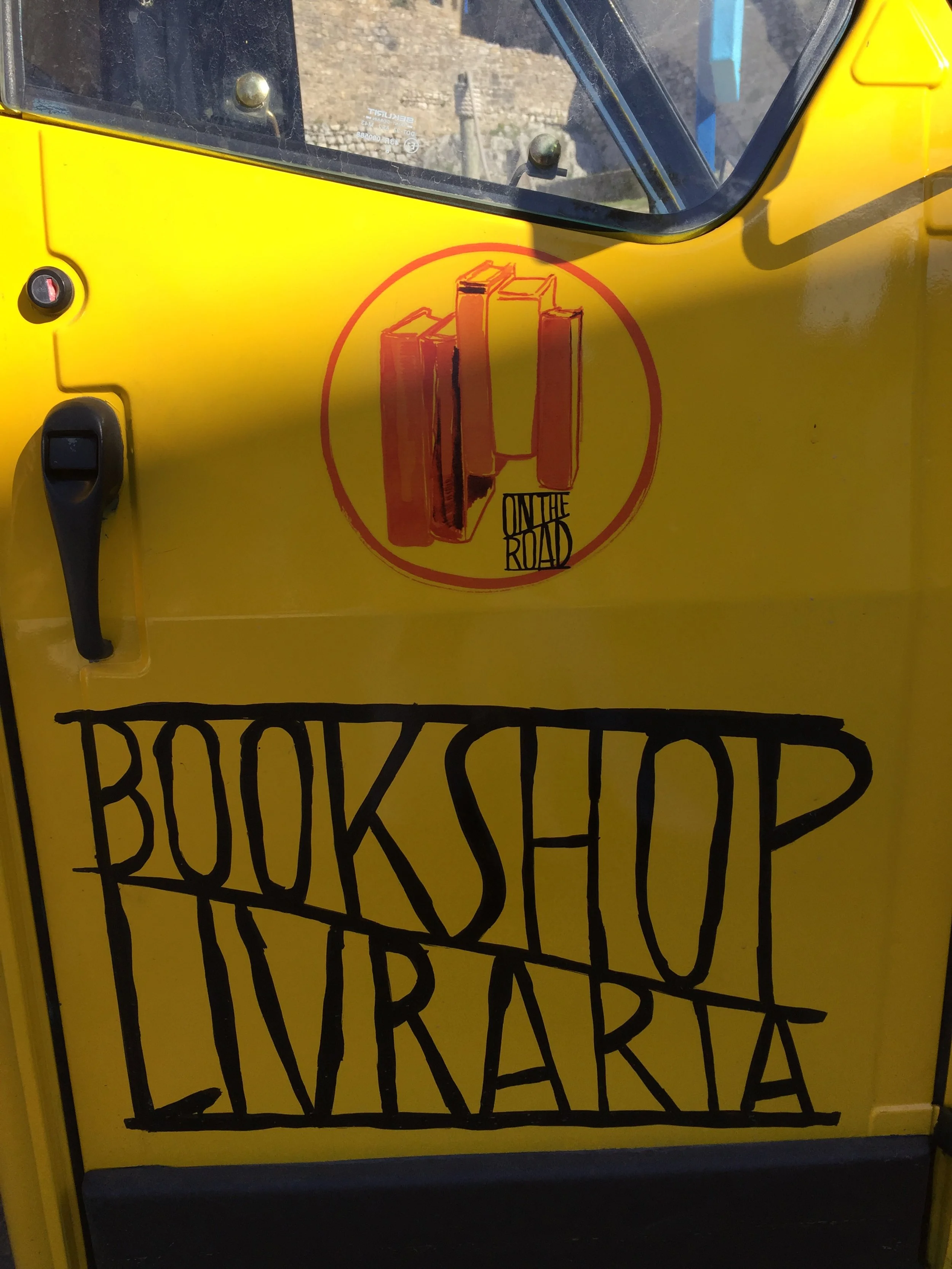 Yellow vehicle with a sticker of a stack of books and the text "On The Road" and handwritten sign "Bookshop Livraria" on the door.