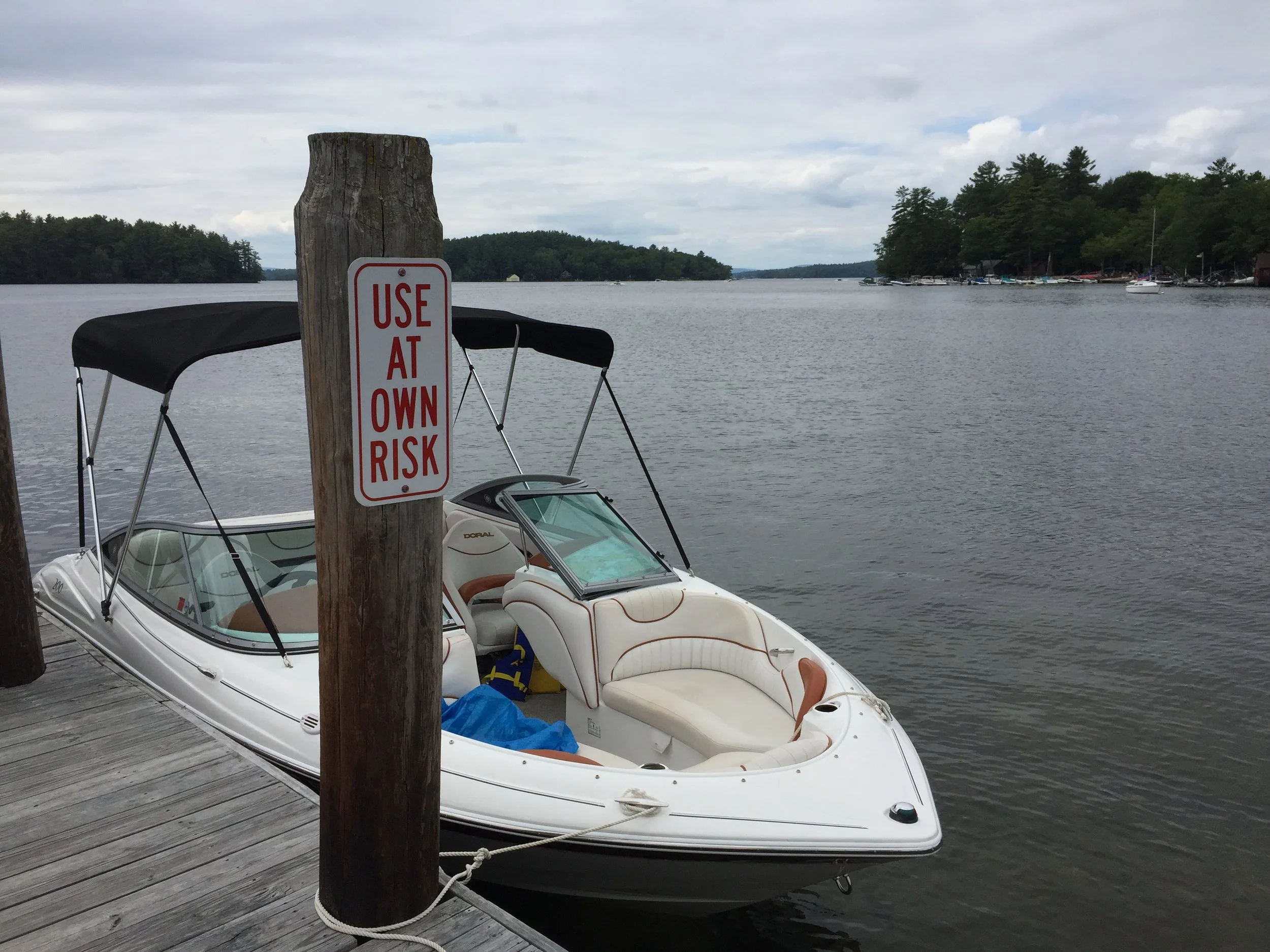 A white motorboat docked at a wooden pier with a sign that reads 'Use at own risk'. The boat has beige seating and is on a calm lake with trees and boats in the background.