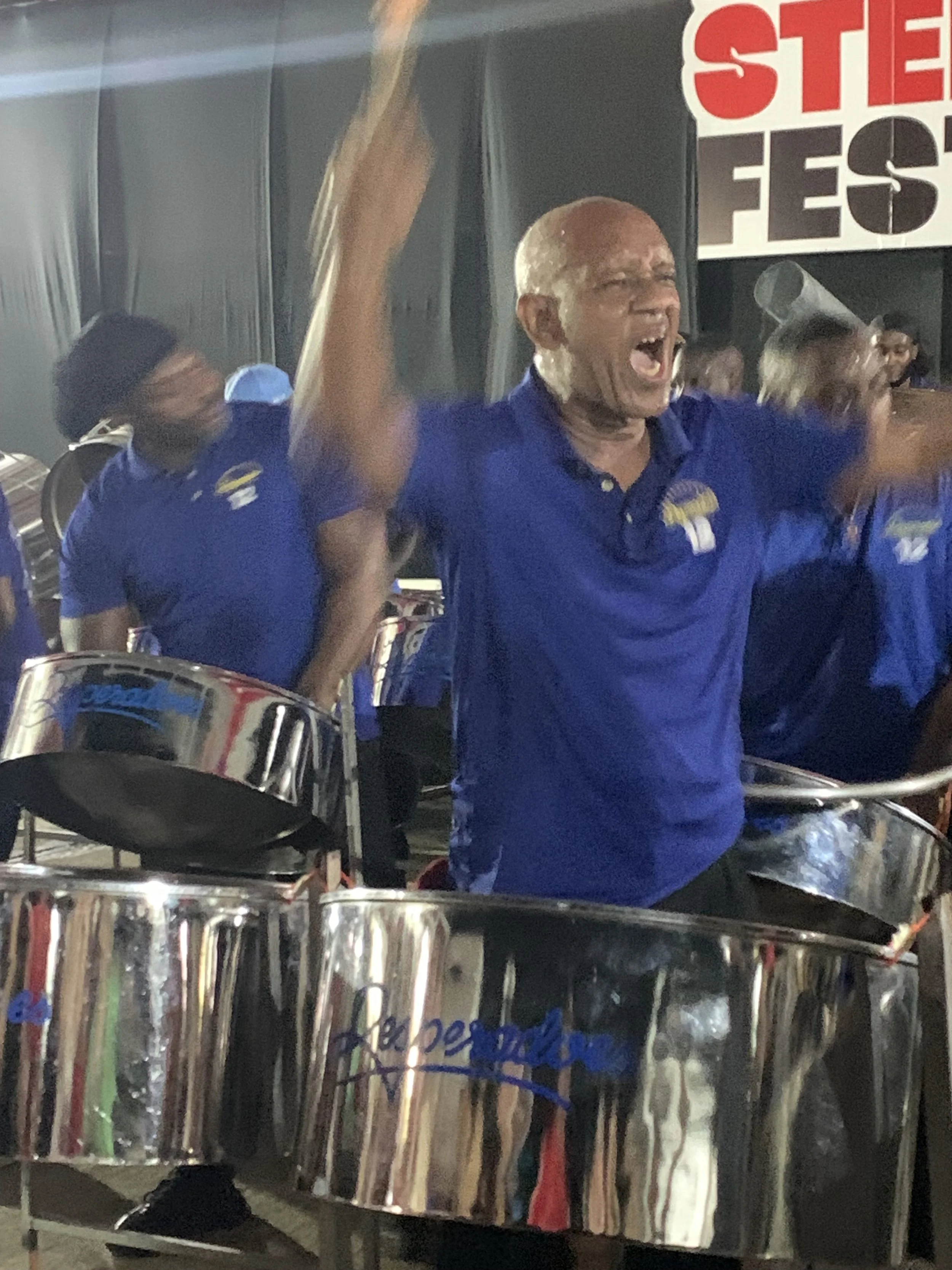A man wearing a blue shirt is playing steel drums and cheering with his hands raised. Several other musicians in blue shirts are in the background, also playing drums. There is a red and white banner that reads "STEE" and "FEST" in the background.