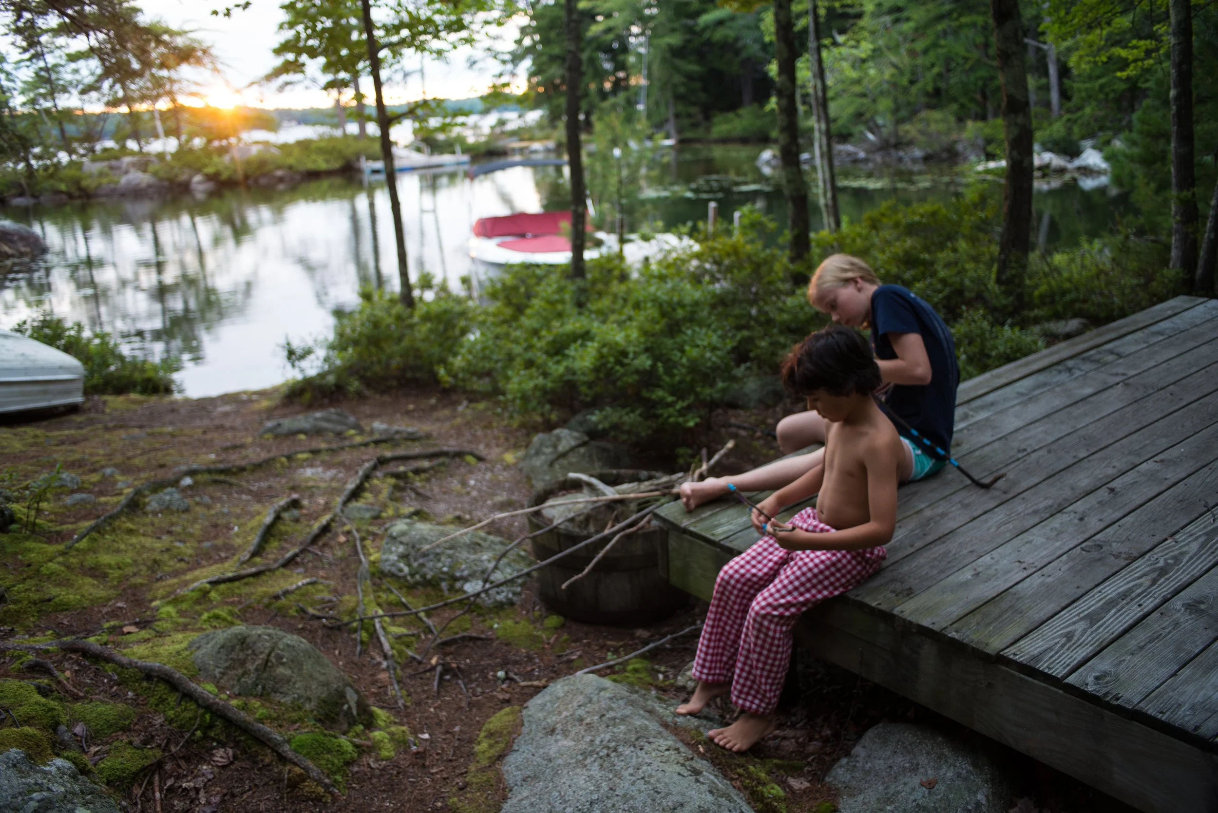 Two children sitting on a wooden dock by a lake at sunset, with several small boats moored nearby and surrounded by trees.