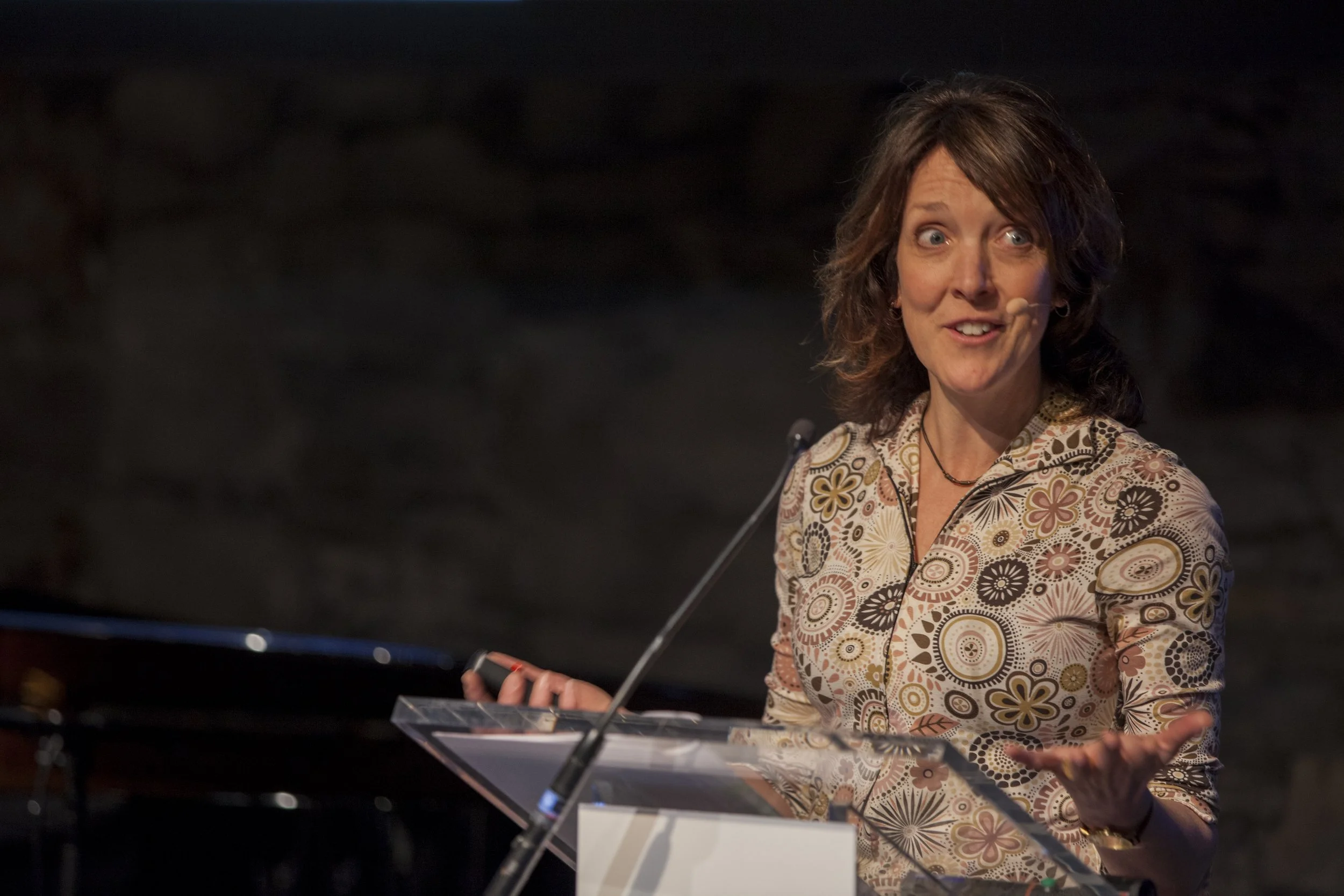A woman with brown, shoulder-length hair speaking at a clear podium, gesturing with her right hand, wearing a patterned blouse, in front of a dark background.
