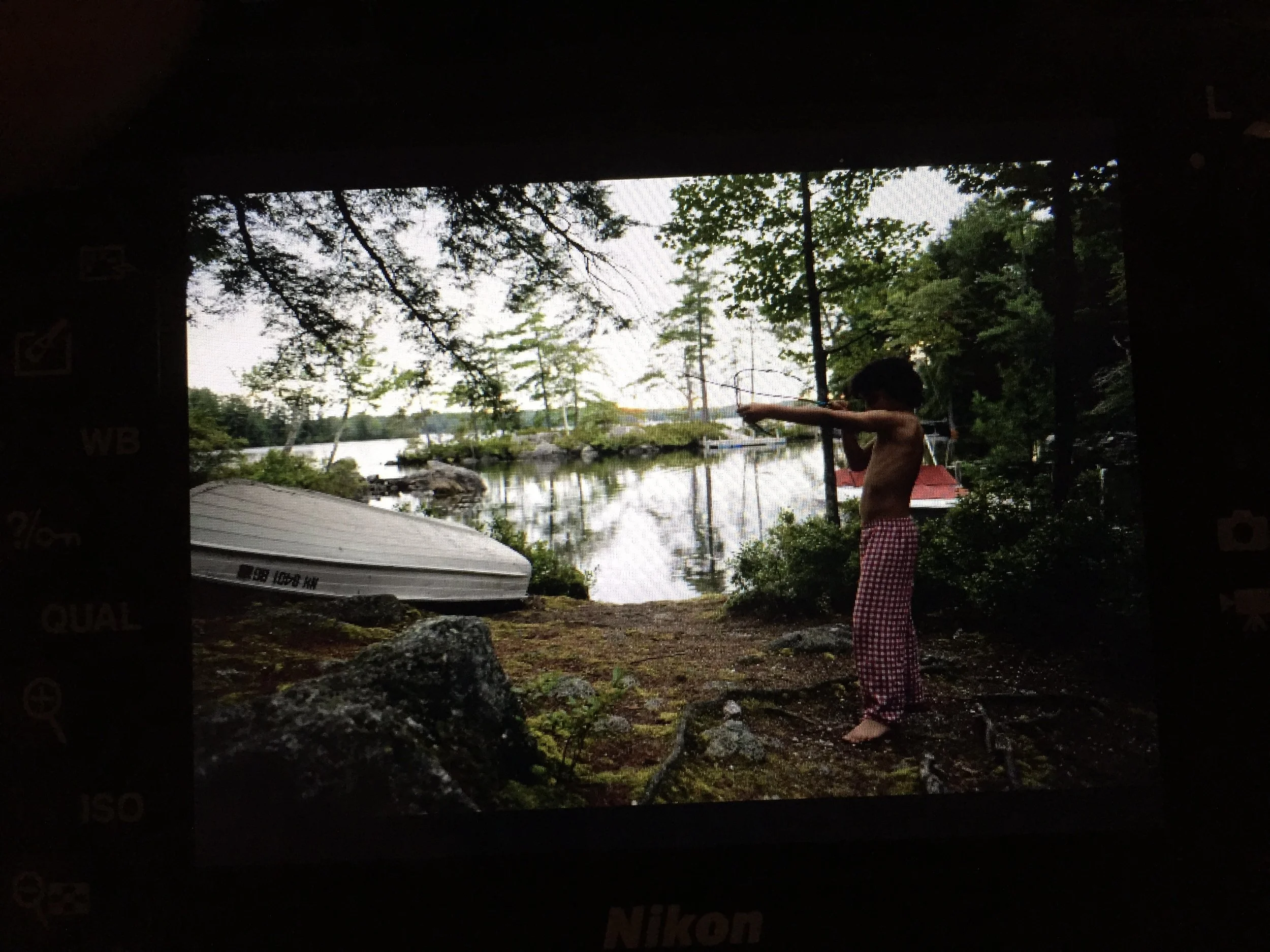 A young boy standing barefoot outdoors near a lake, stretching his arms forward, surrounded by trees and rocks, with a boat and other boats in the background.