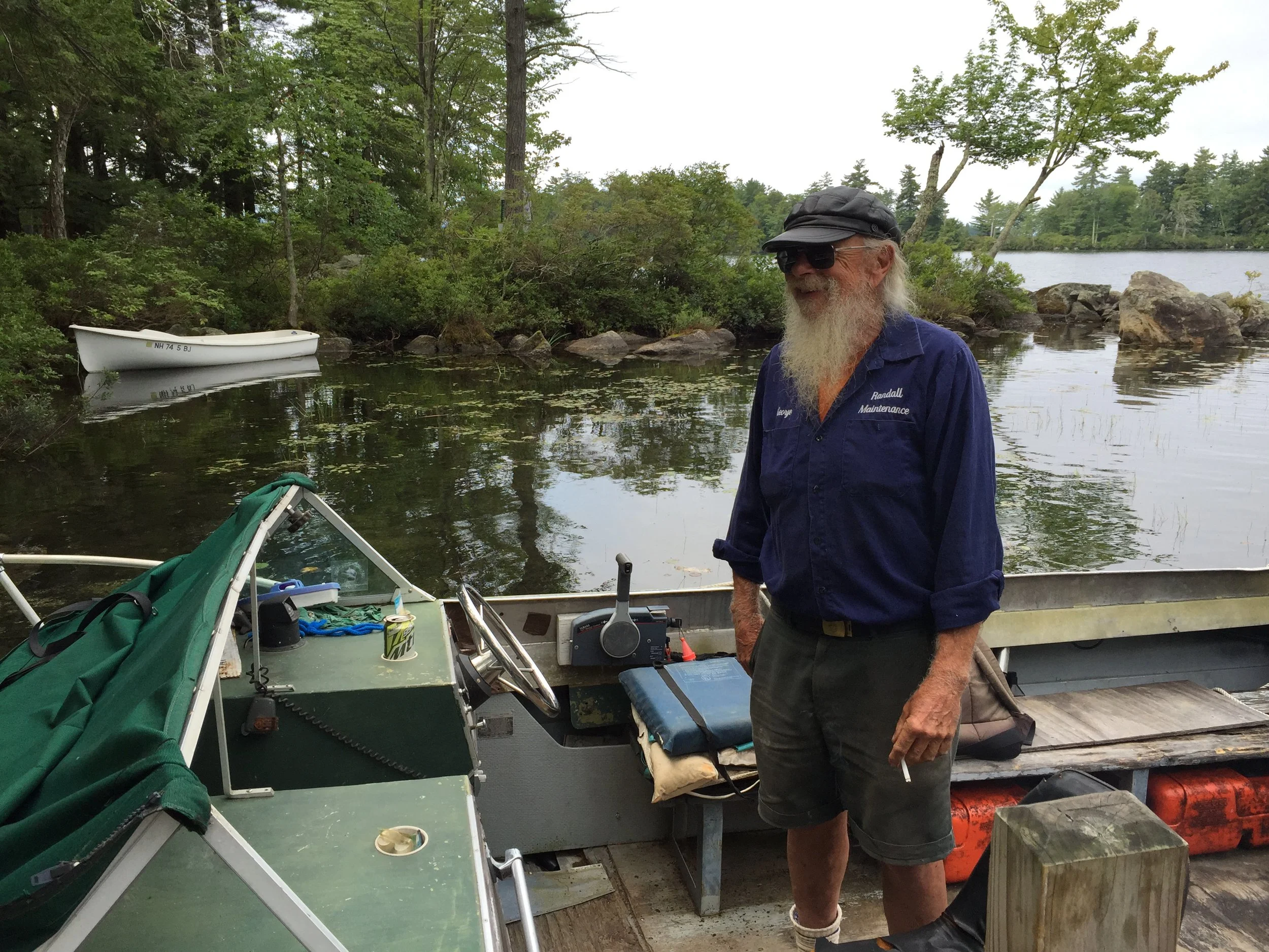 A man with gray hair and a beard standing on a boat dock near a boat, with a lake and trees in the background.