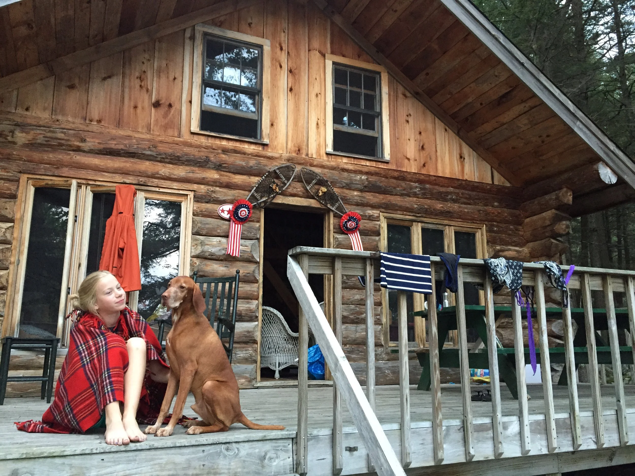 A young girl and a dog sitting on a wooden porch of a log cabin, with laundry hanging on the railing, and an axe decoration with red, white, and blue ribbons on the wall.