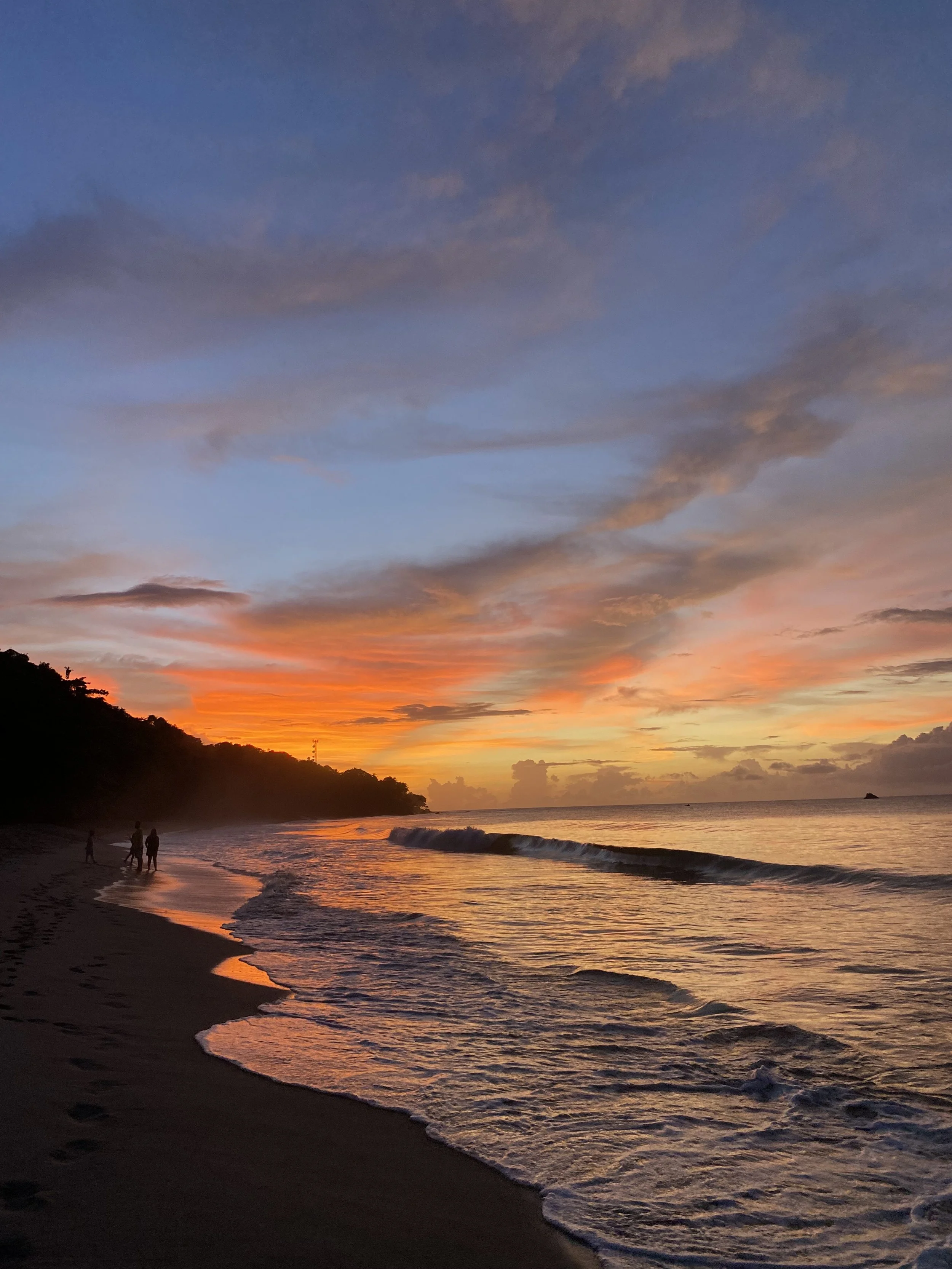 Sunset over the ocean with colorful sky, shoreline, and people walking on the beach.