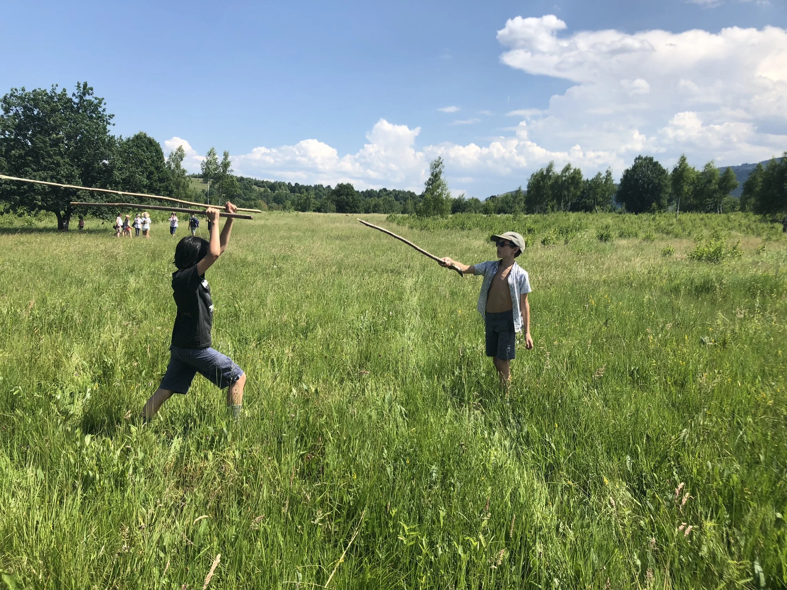 Two boys are engaged in a mock sword fight with sticks in a grassy field, with a group of people in the background under a partly cloudy sky.