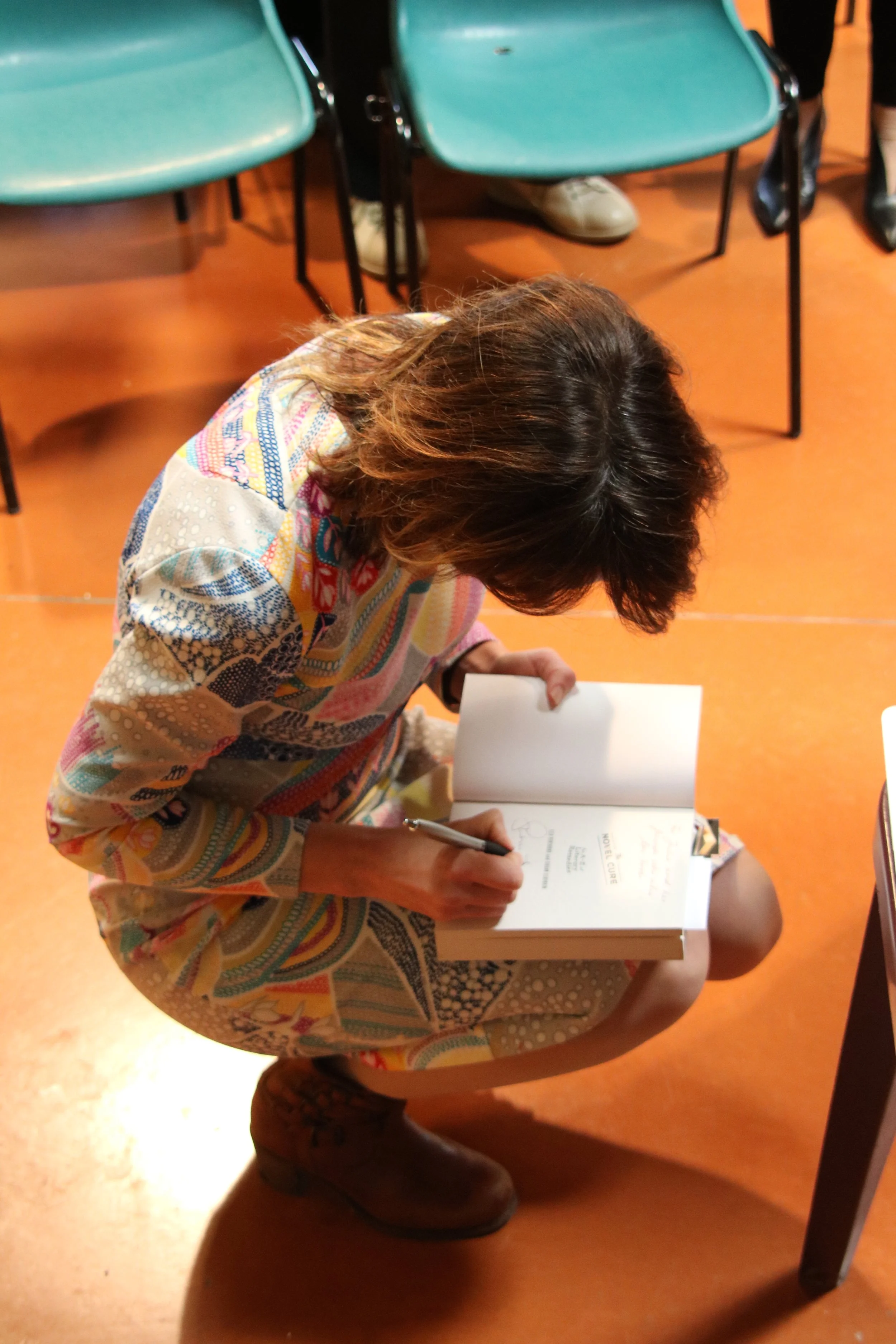 A woman with brown hair, wearing a colorful patterned dress and brown boots, is sitting on the floor and writing in a book.
