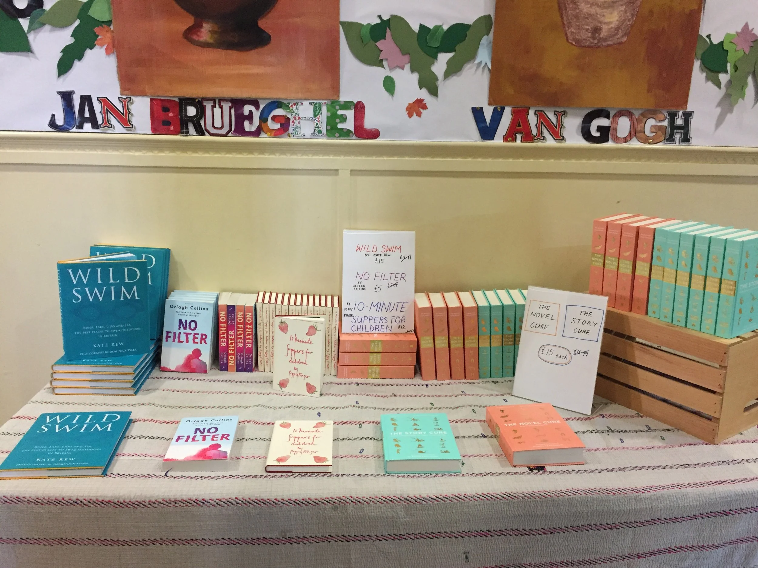 Display of books by Kate Rew at a book sale, including titles 'Wild Swim' and 'No Filter'. Signage indicates prices and book series 'The Novel Cure' and 'The Story Cure'. Background features a banner with colorful cutouts and the text 'Jan BrugeL Van