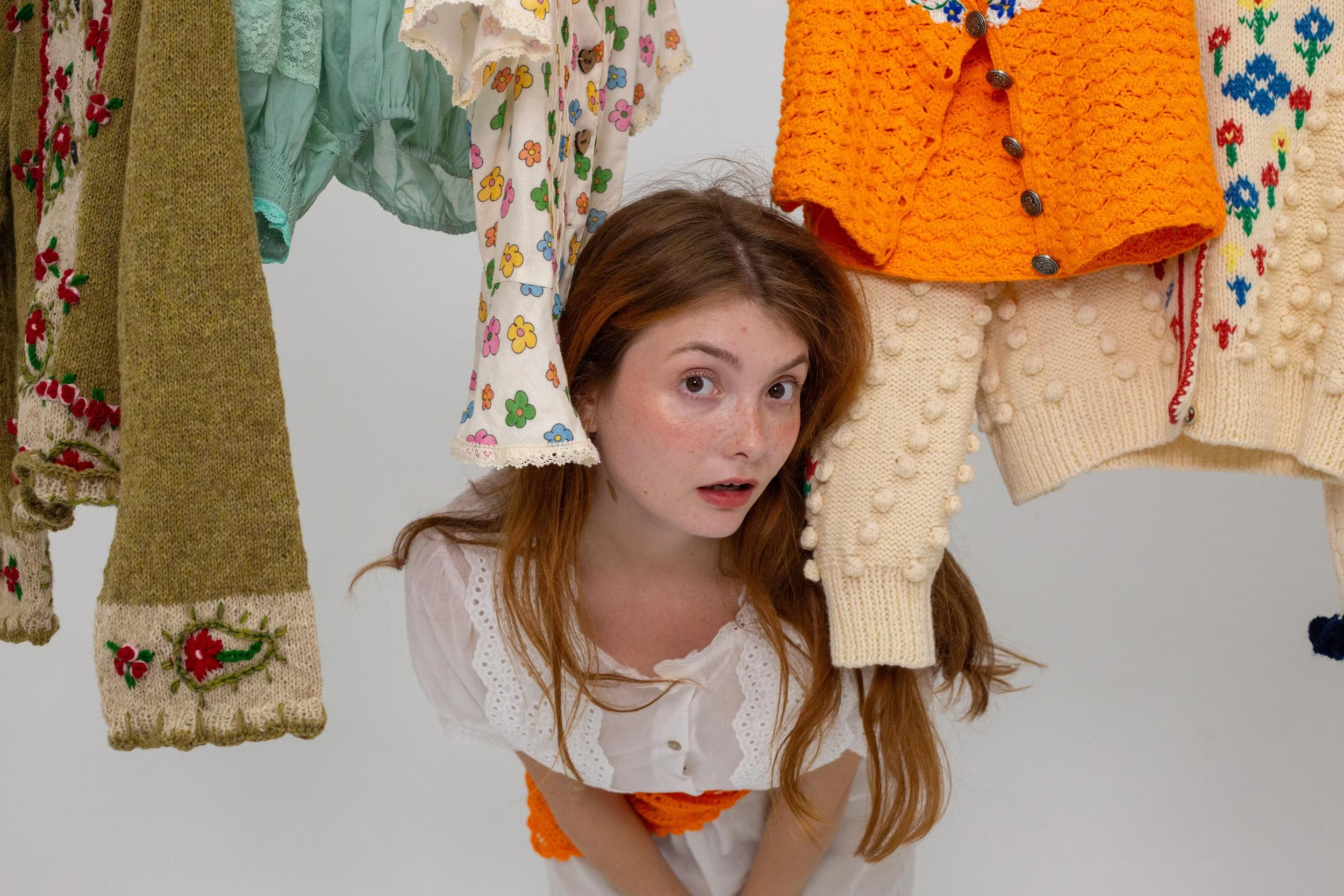 Une jeune femme aux cheveux roux, portant une robe blanche avec broderies, regarde vers le haut à travers des vêtements colorés suspendus.