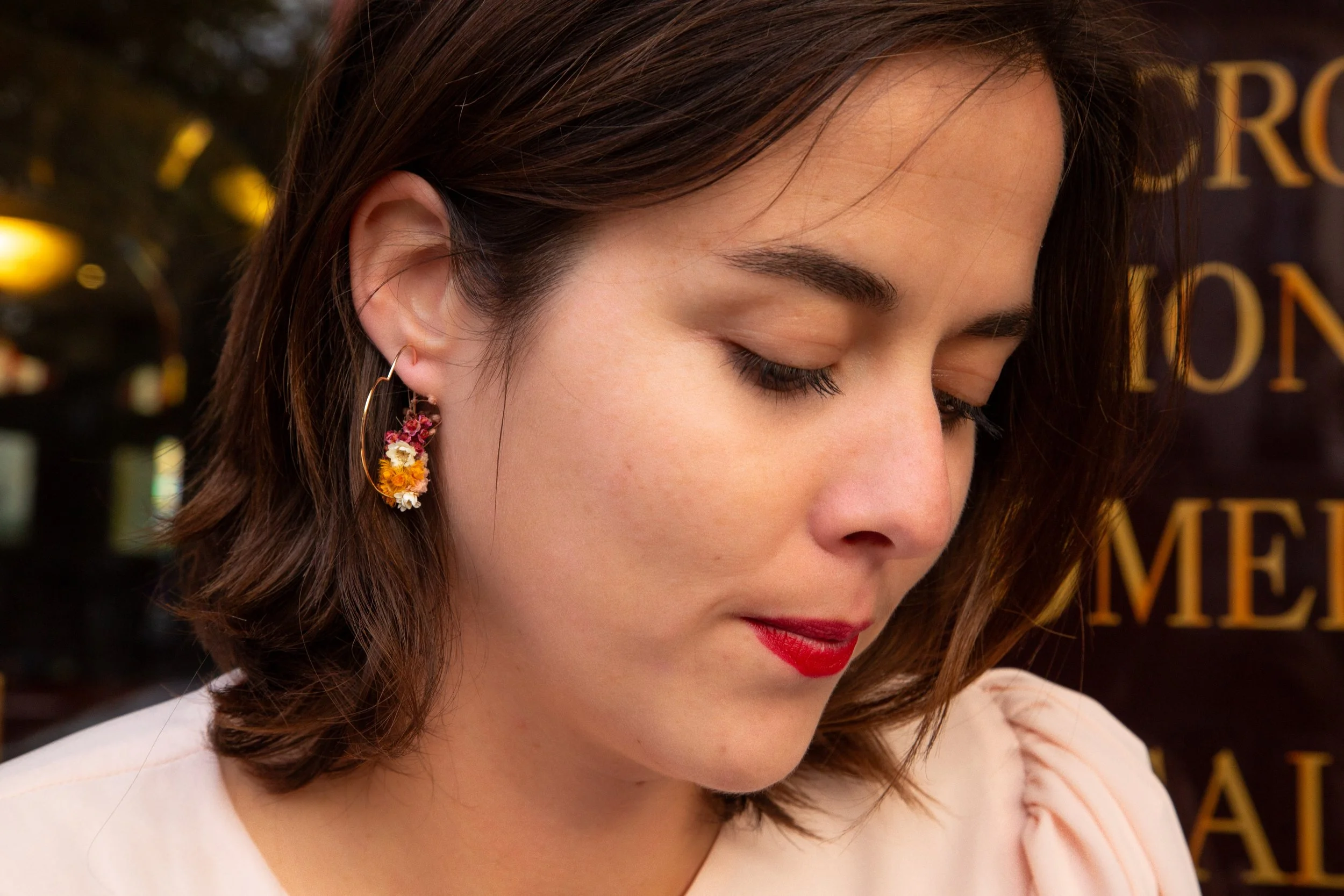 Jeune femme aux cheveux bruns, portant une boucle d'oreille florale et un maquillage léger avec un rouge à lèvres rouge, regardant vers le bas, avec un fond flou de texte en français.