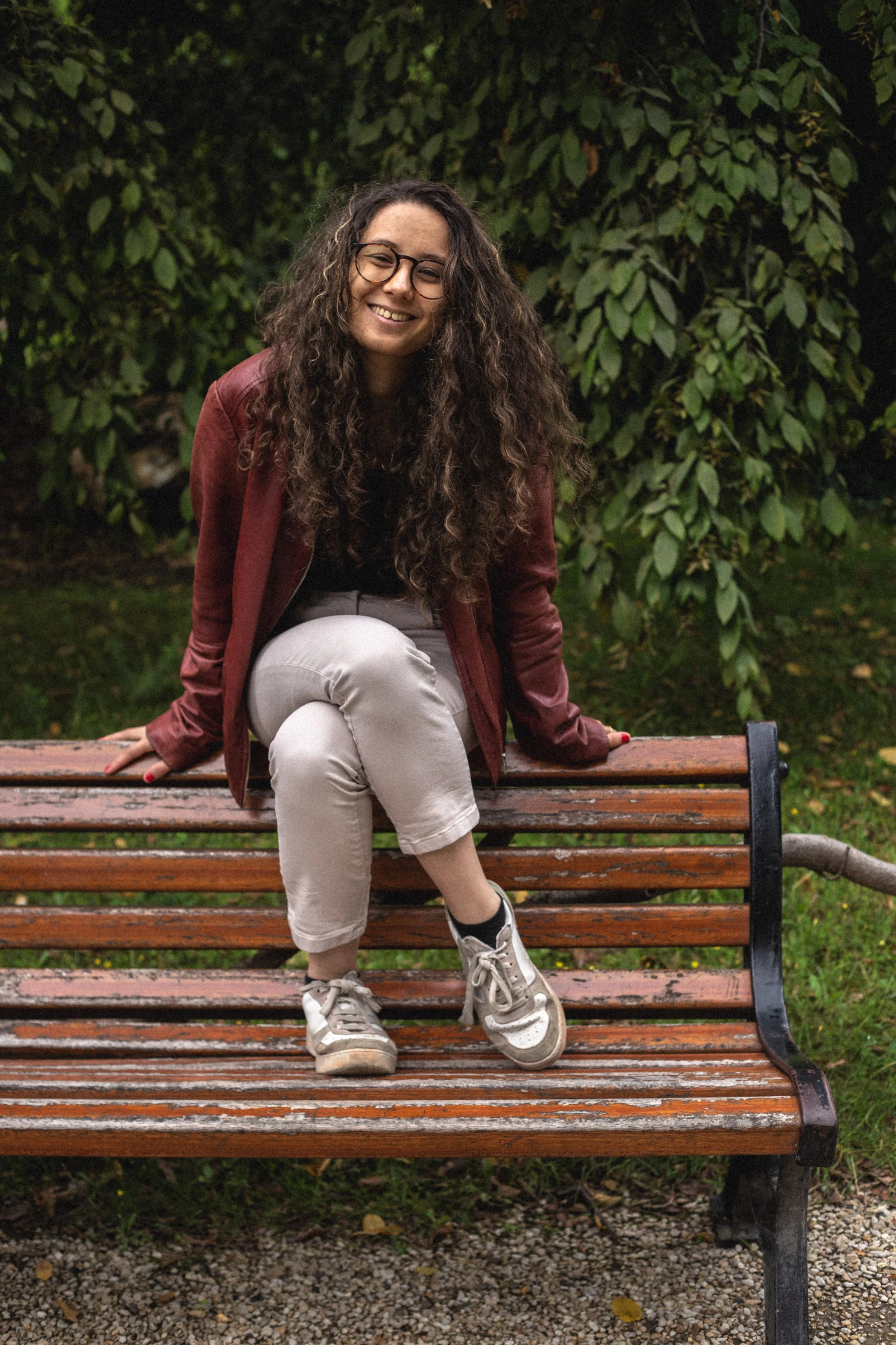 Une jeune femme aux cheveux bouclés assise sur un banc en bois dans un parc, souriant à la caméra.