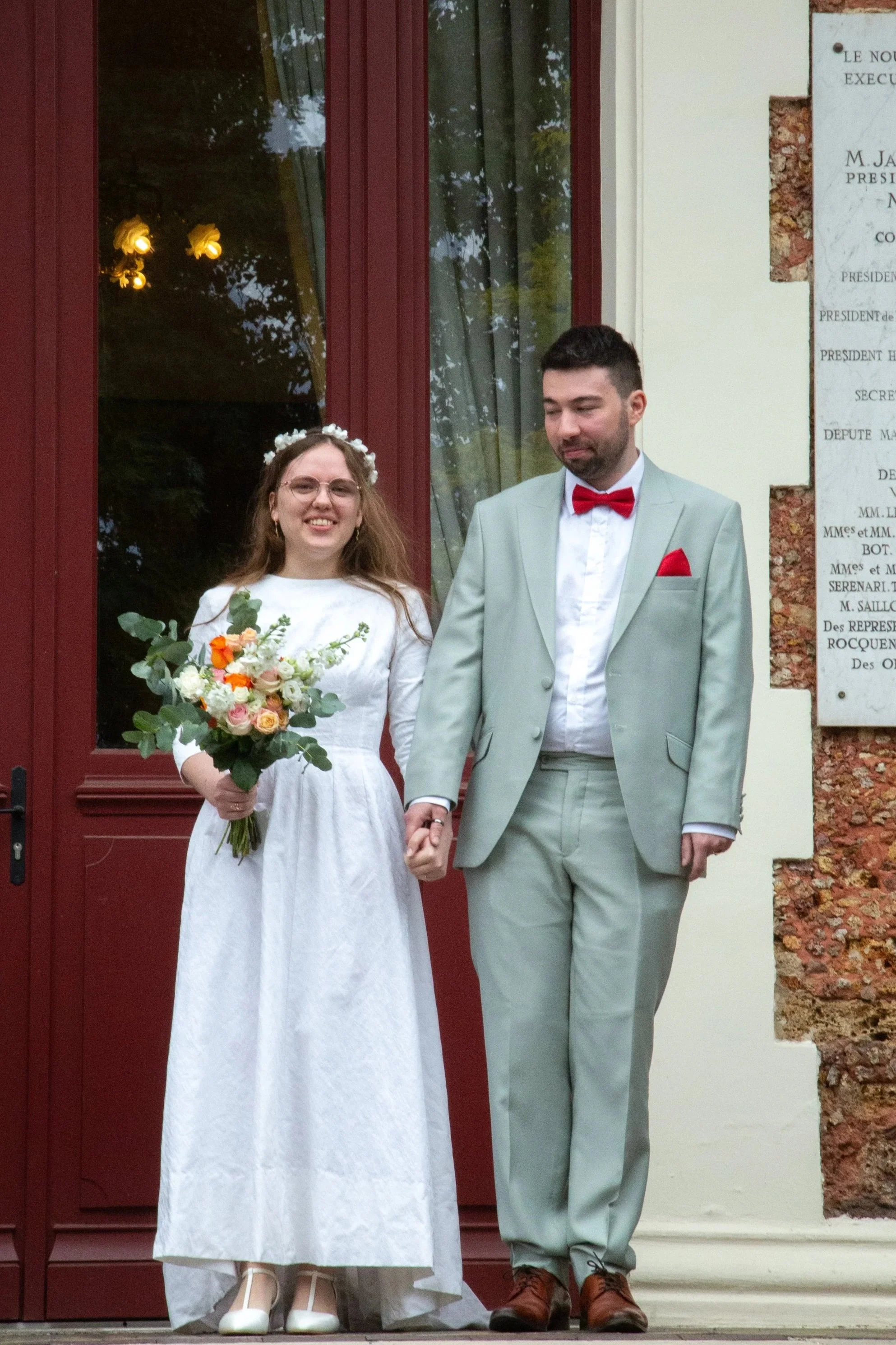Un couple de mariés, une femme en robe blanche tenant un bouquet de fleurs et un homme en costume gris avec une cravate corail, se tenant par la main devant une porte rouge pendant leur mariage.
