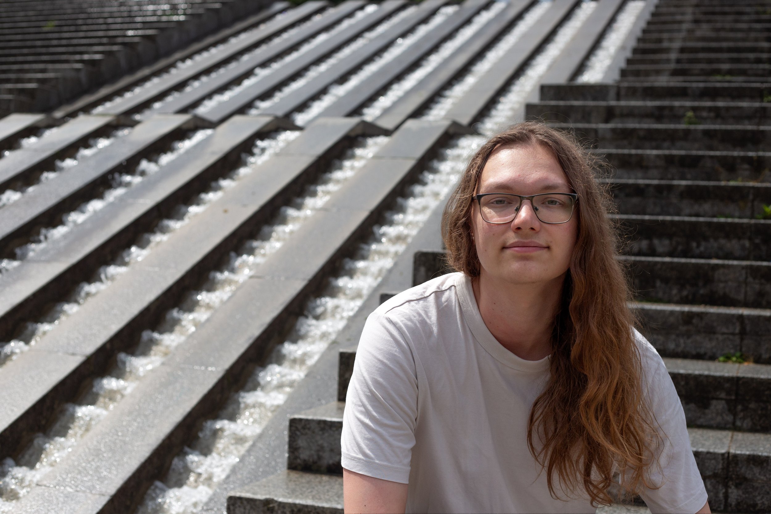 Un homme avec des lunettes et des cheveux longs assise devant un escalier en béton avec une fontaine d'eau passant entre les marches.