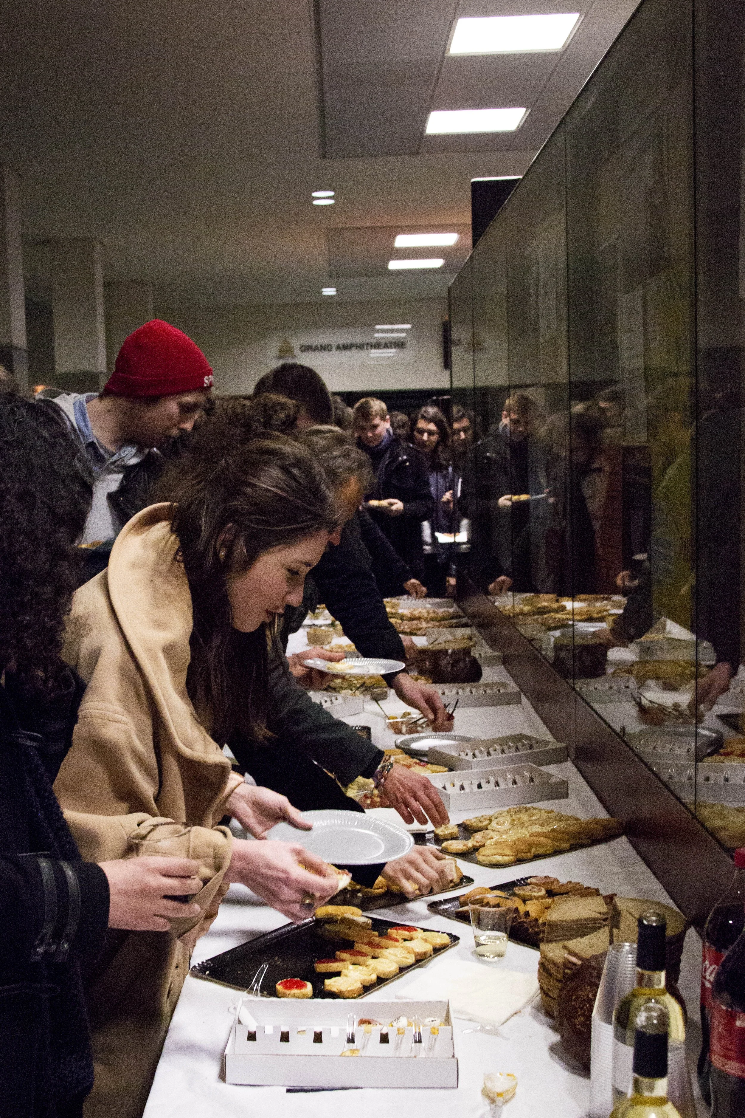 Des personnes font la queue pour se servir lors d'un buffet de desserts dans une cafétéria, avec un panneau indiquant « Grand Amphithéâtre » en arrière-plan.