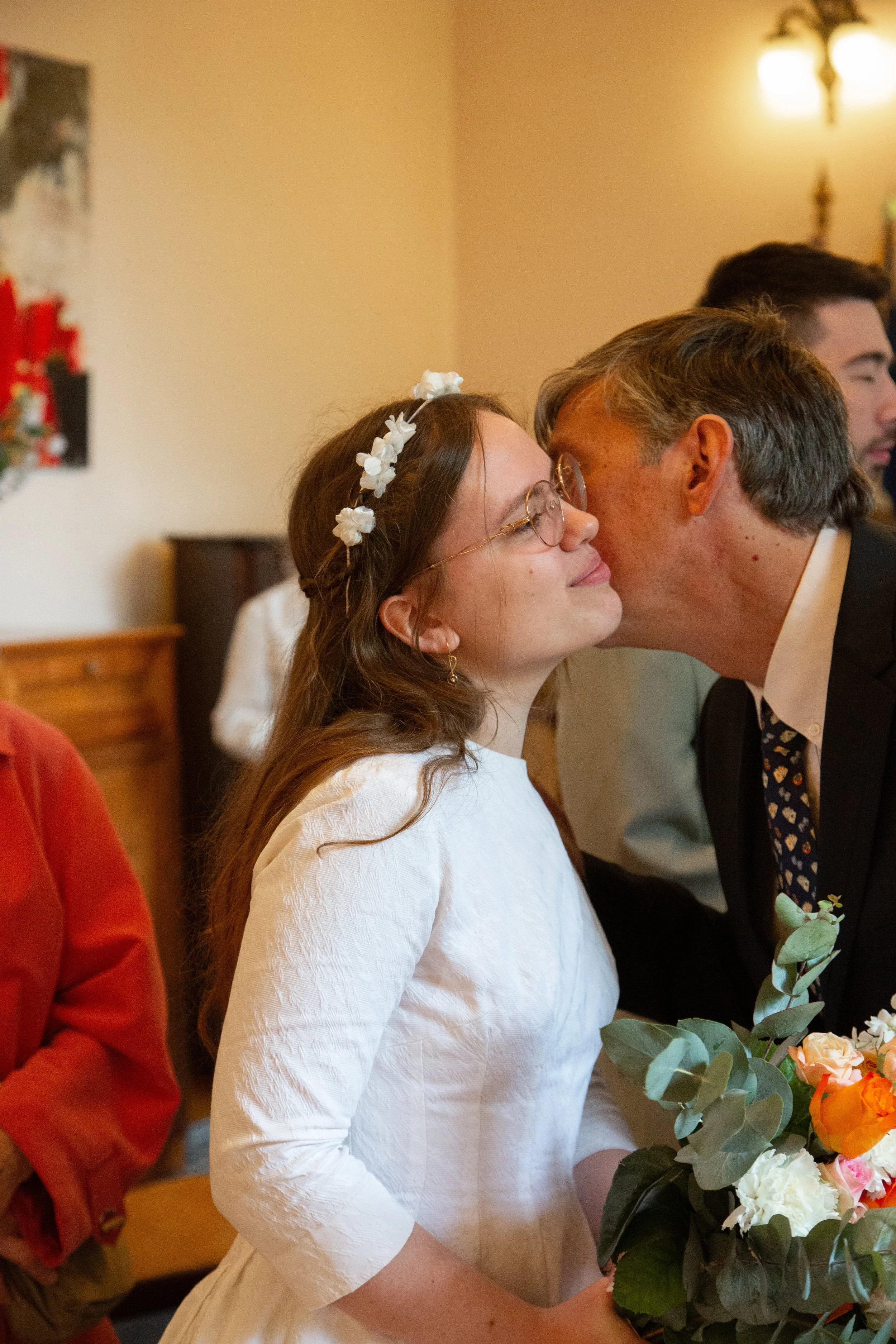 Une jeune femme en robe blanche et avec une couronne de fleurs reçoit un baiser sur la joue d'un homme en costume lors de son mariage.