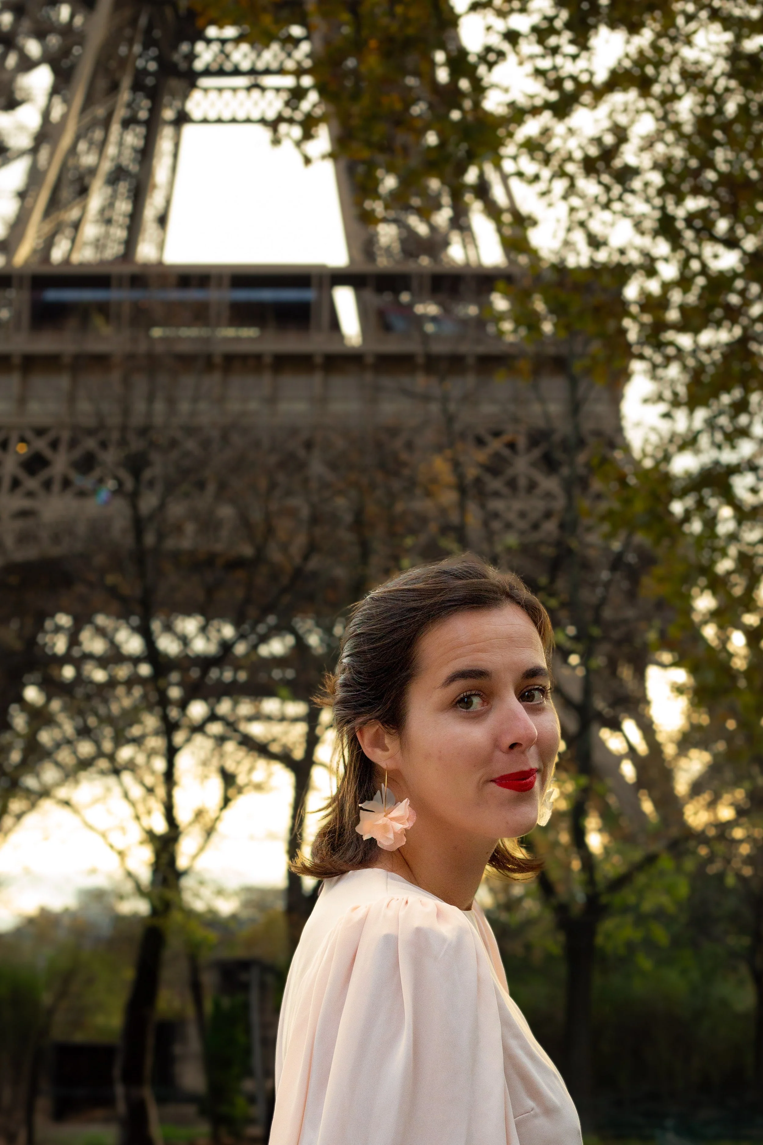 Une femme souriante avec des boucles d'oreilles en fleurs, portant une robe claire, se tenant à l'extérieur près de l'Éiffel Tower avec des arbres en arrière-plan.